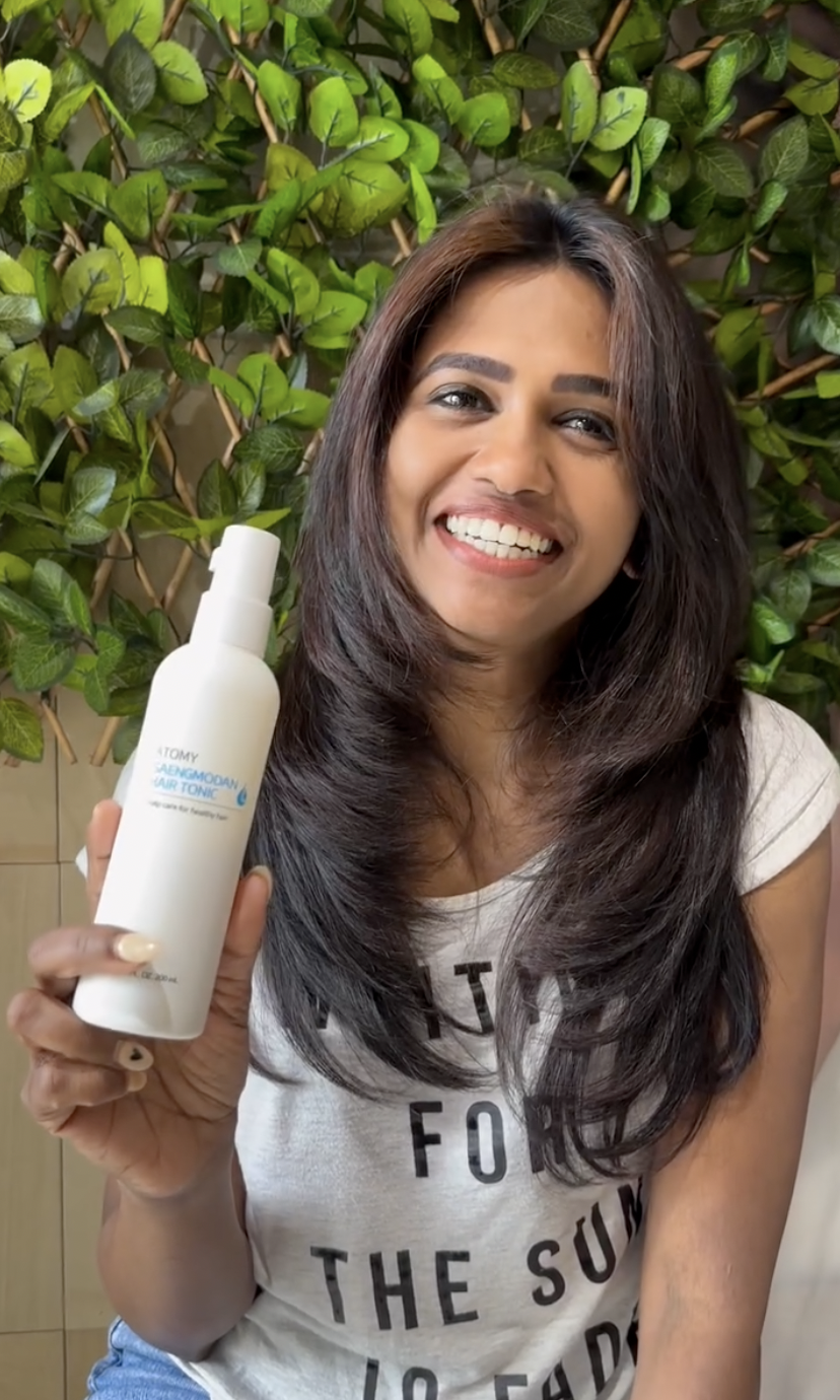 A smiling woman with long dark brown hair holding a white spray bottle labeled "TONY" in front of a background of green leafy plants.