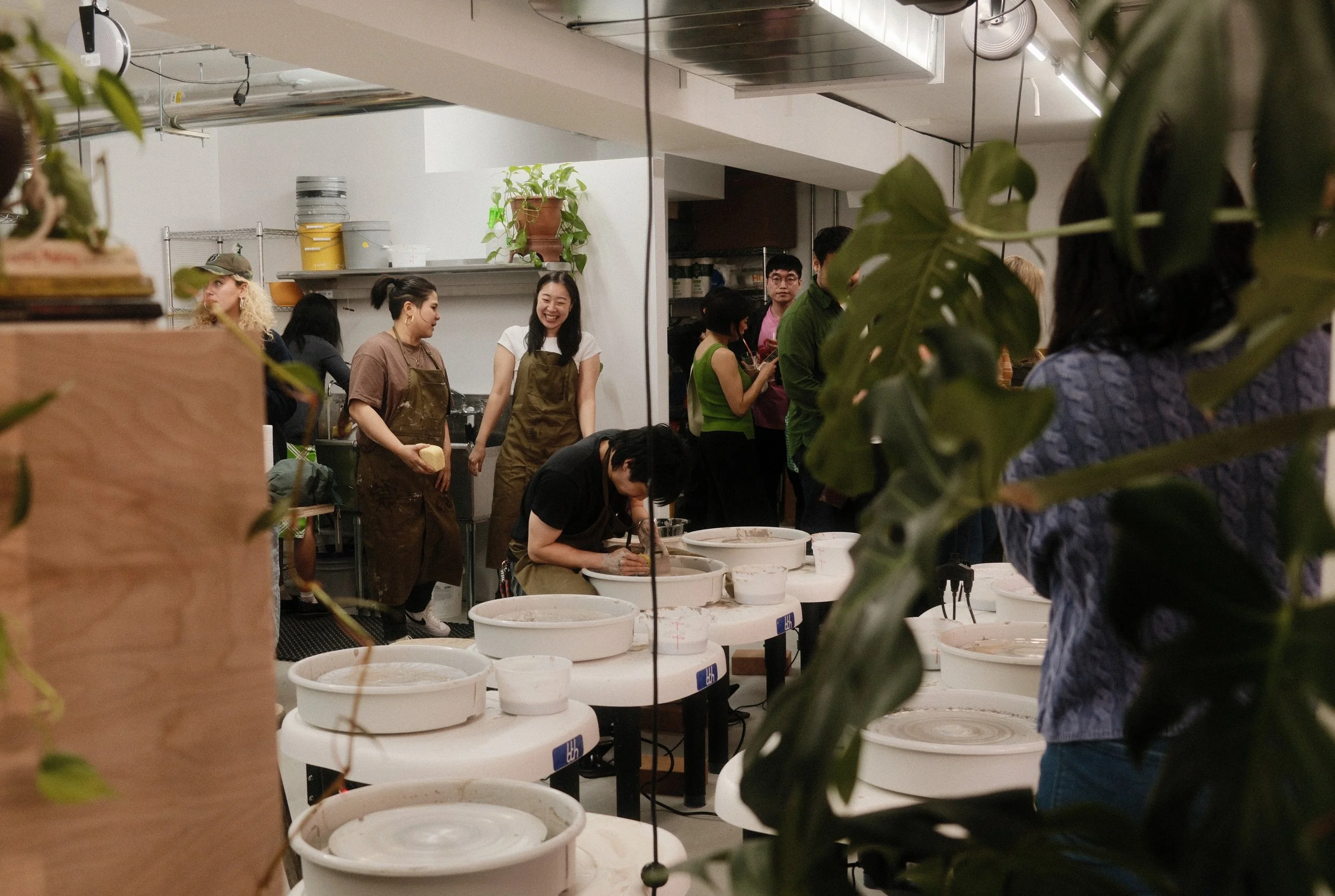 A pottery studio with a male student shaping clay on a potter’s wheel while two women in aprons talk nearby. A crowd of people is visible in the background, and green plants line the sides of the studio.