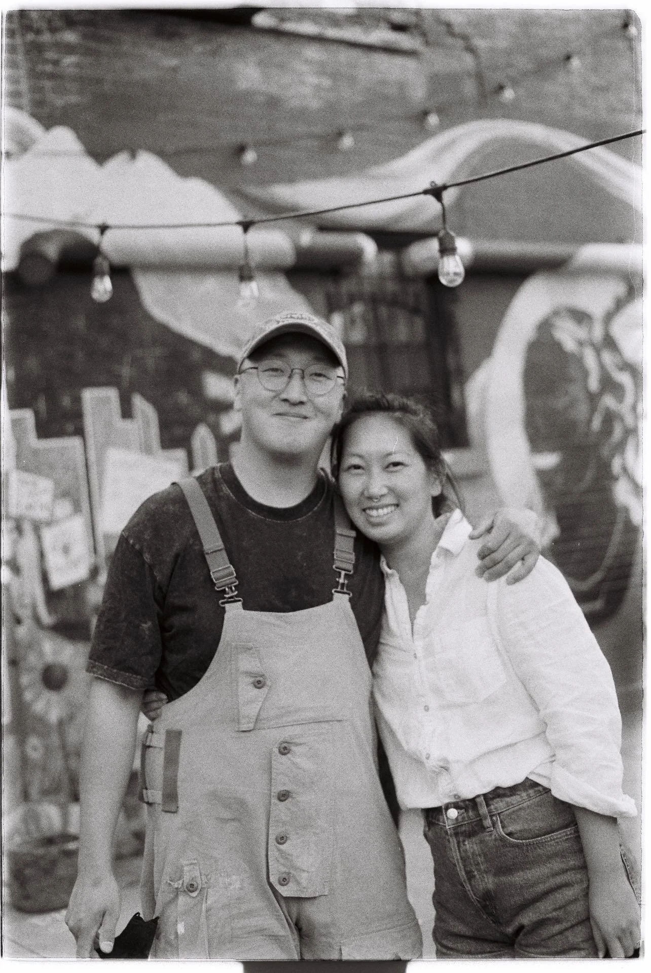 Black-and-white photo of a man wearing overalls, a hat, and round-frame glasses, with his arm around a woman. She is wearing a white linen long-sleeve top and blue jeans. They are standing in front of a mural, with string lights overhead.