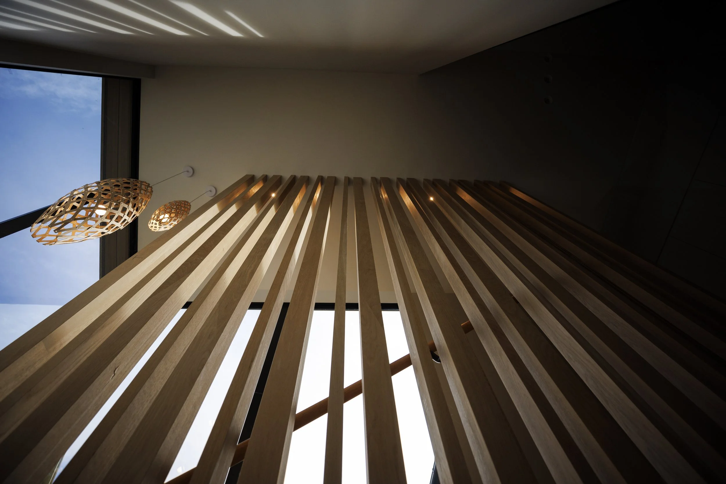 Interior view of a modern space with feature vertical wooden slats to a stairwell, a Trubridge pendant light, and a window at the landing with a clear blue sky.