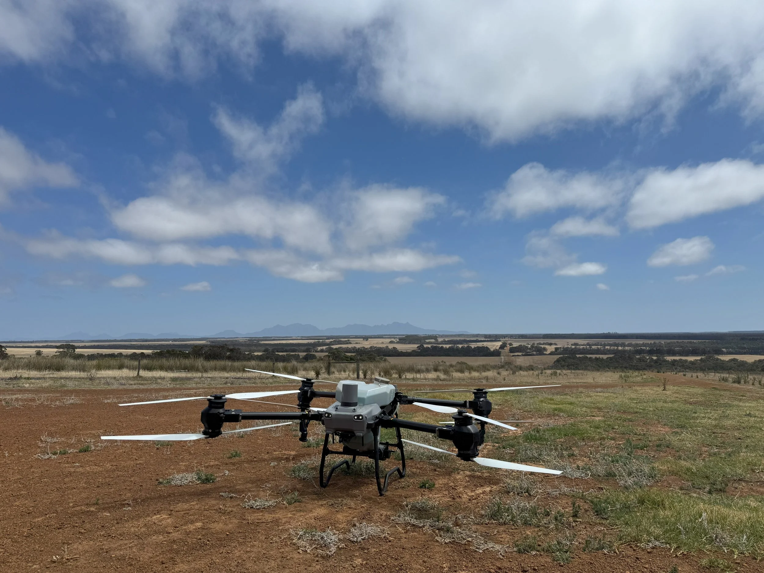 A drone with multiple rotors on open land with dry grass and shrubs, under a partly cloudy blue sky.
