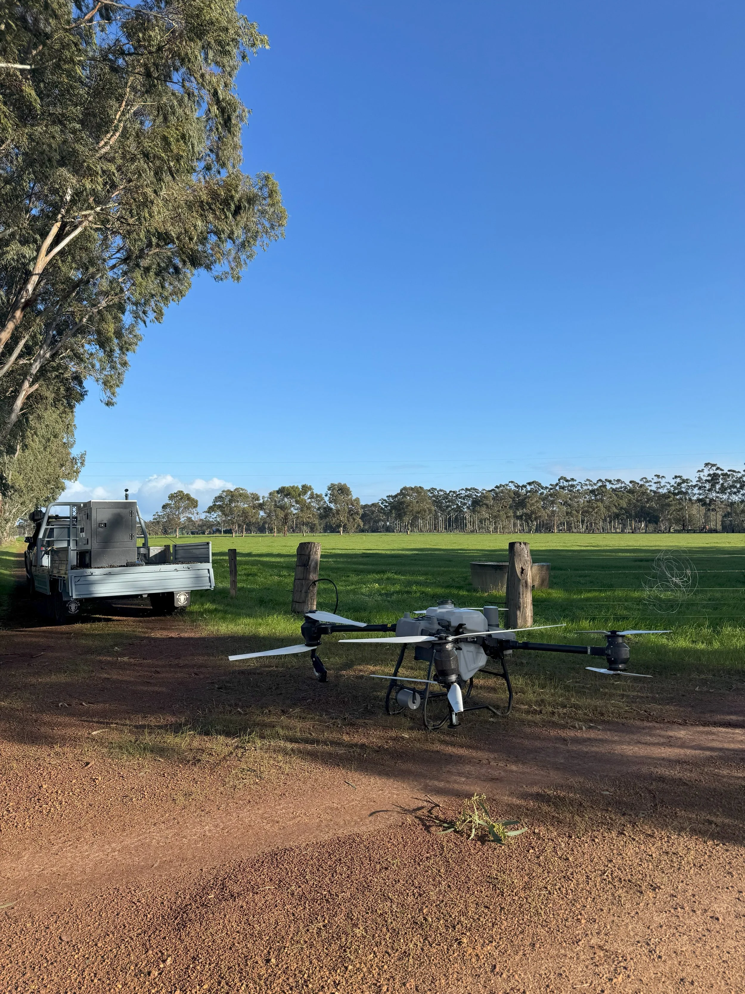 A drone and a truck parked on a dirt path beside a grassy field with trees and a clear blue sky in the background.