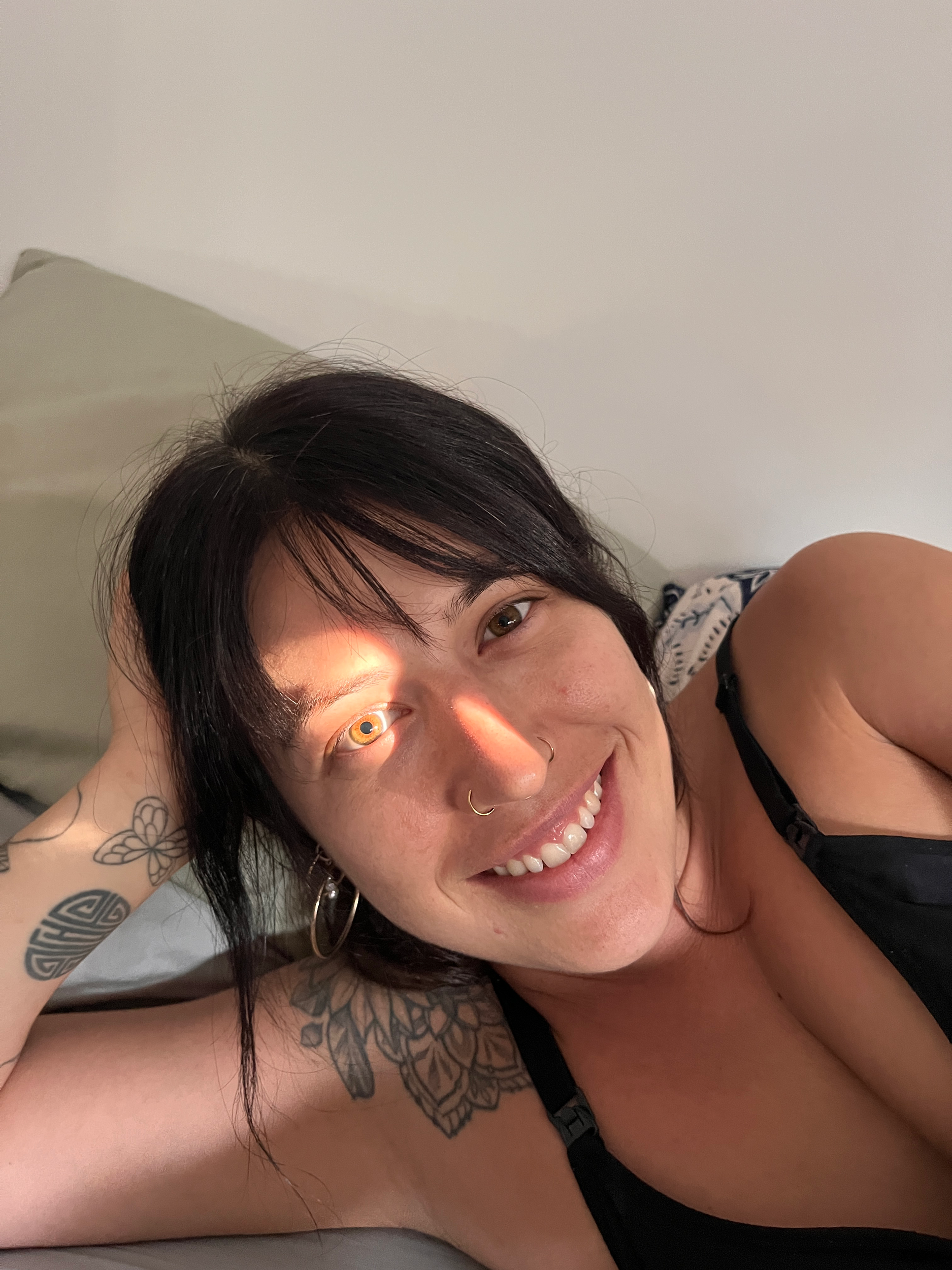 Close-up of a smiling woman with black hair, tattoos on her arm, and wearing black clothing, lying down with her head resting on a pillow.