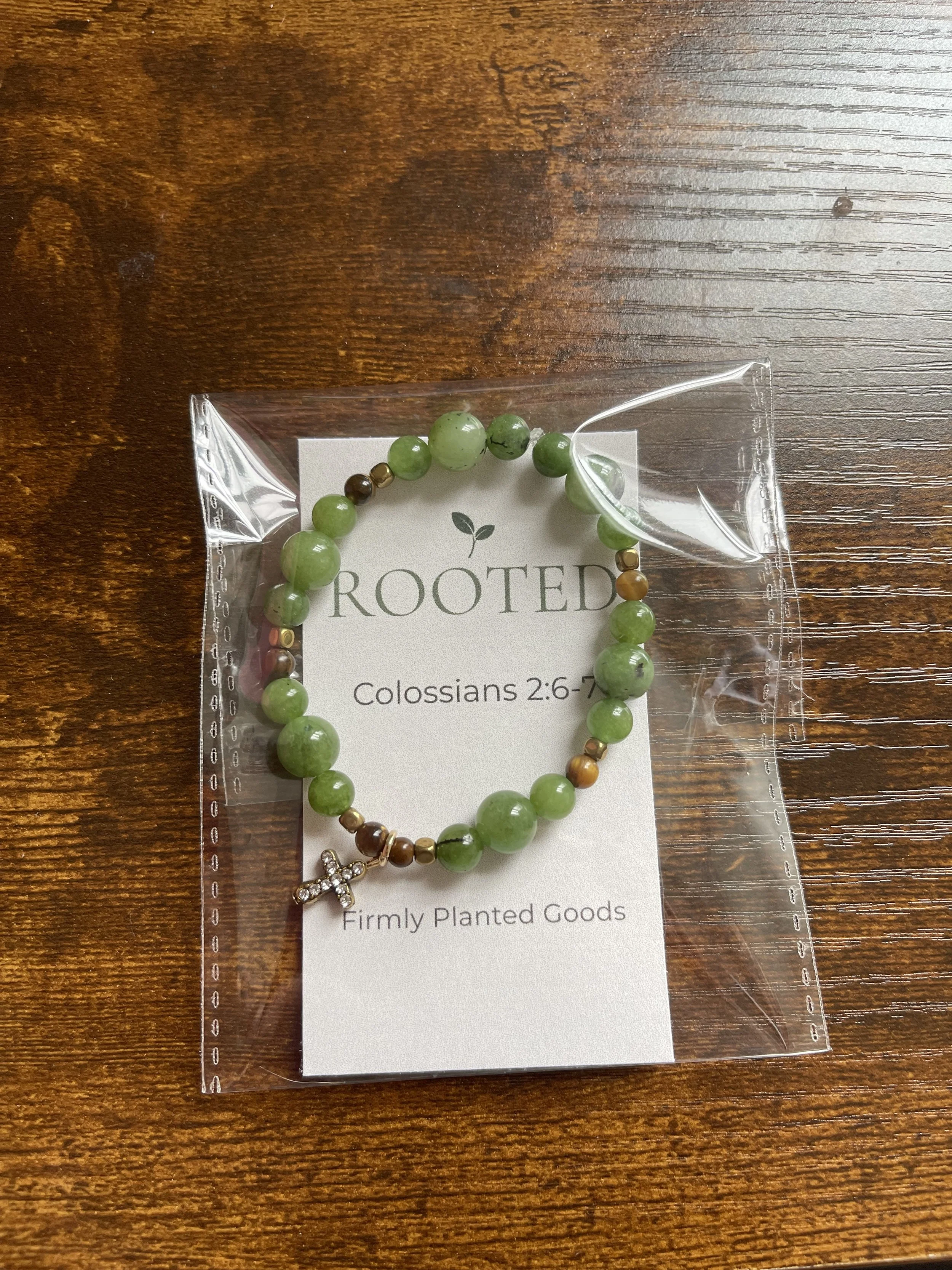 Green beaded bracelet with a cross charm, packaged on a card that says 'ROOTED' and references Colossians 2:6-7, on a wooden surface.
