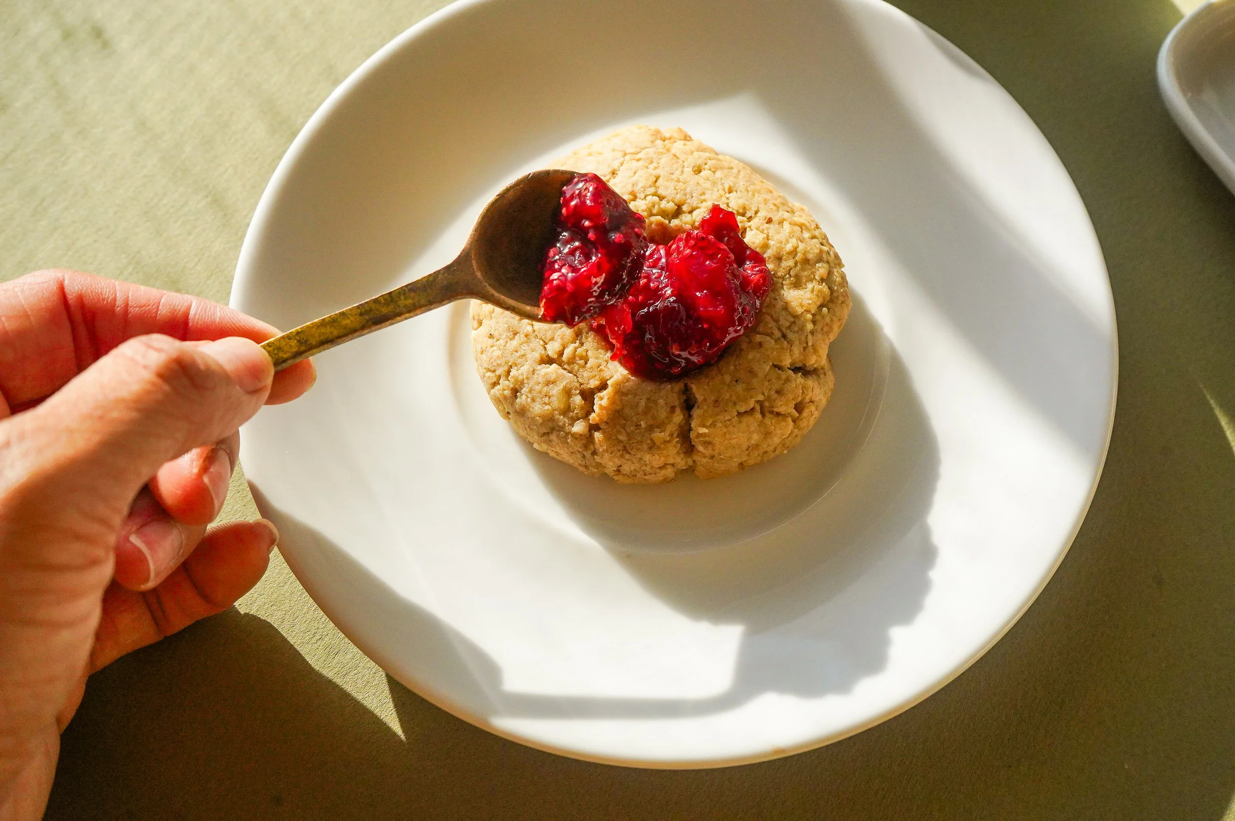 A treat being served on a white plate, featuring a large, round, crumbly thumbprint cookie topped with a spoonful of red berry jam or compote from The Delicate Squash, with a person's hand holding a gold antique spoon over it.