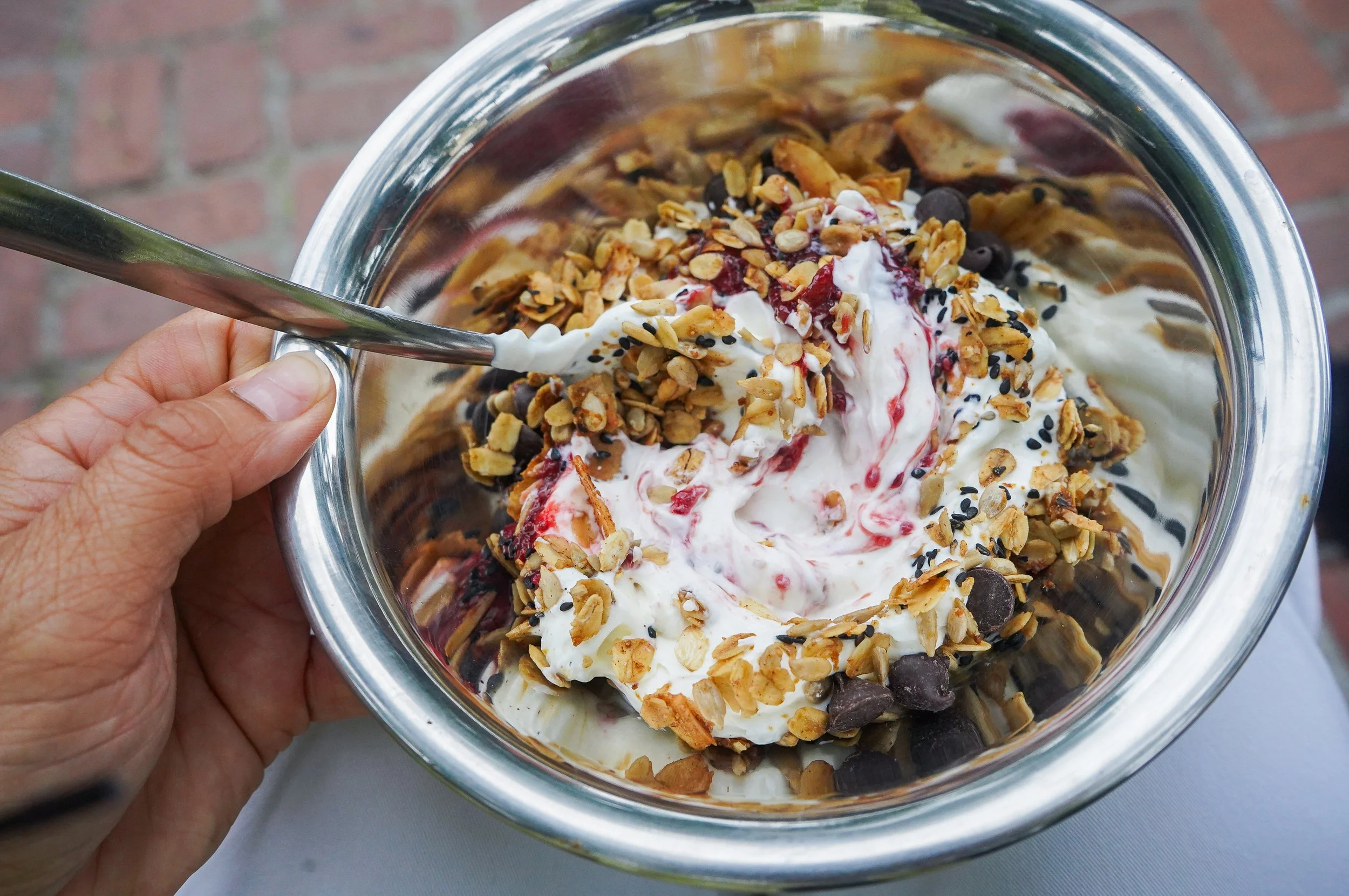 A hand holding a metal spoon over a bowl of yogurt topped with granola from The Delicate Squash, chocolate chips, and homemade jam.