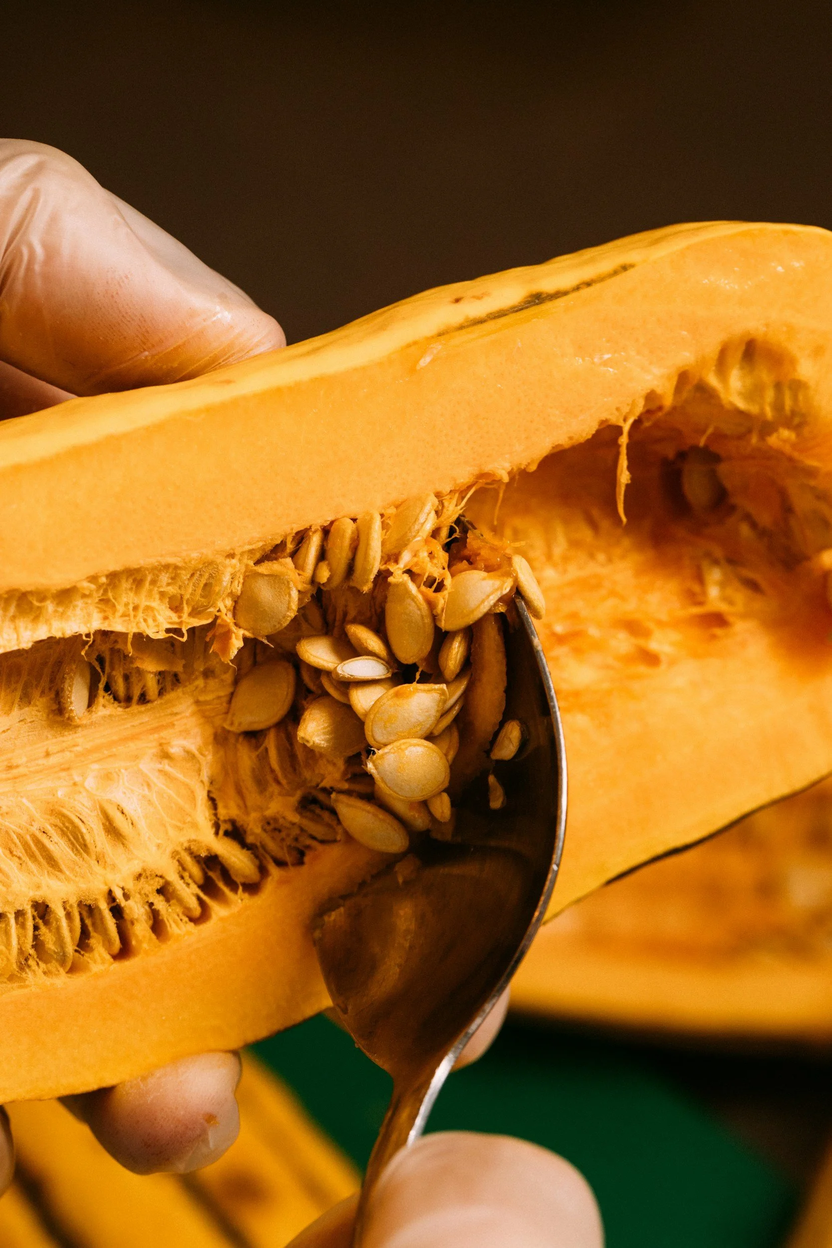 Close-up of a person's hand holding a spoon and scooping out seeds from the orange flesh of a squash.