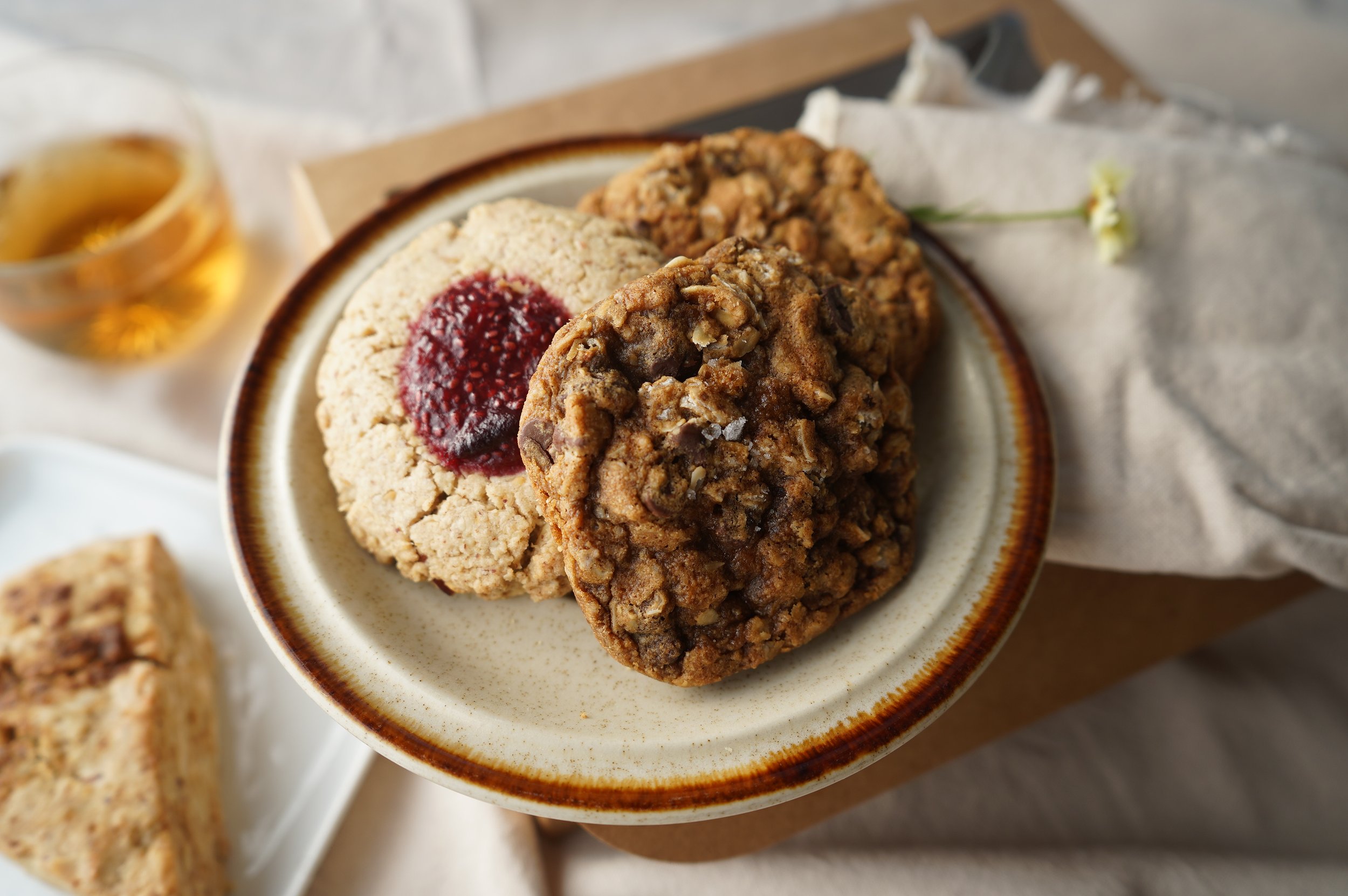 Close-up of two chocolate chip oatmeal cookies and one thumbprint cookie from The Delicate Squash on a vintage plate and a short glass of herbal tea in the background.