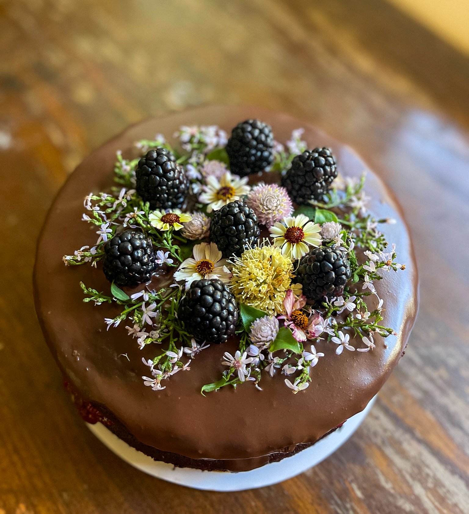 Chocolate cake topped with blackberries and assorted small edible flowers on a wooden surface from The Delicate Squash in San Diego