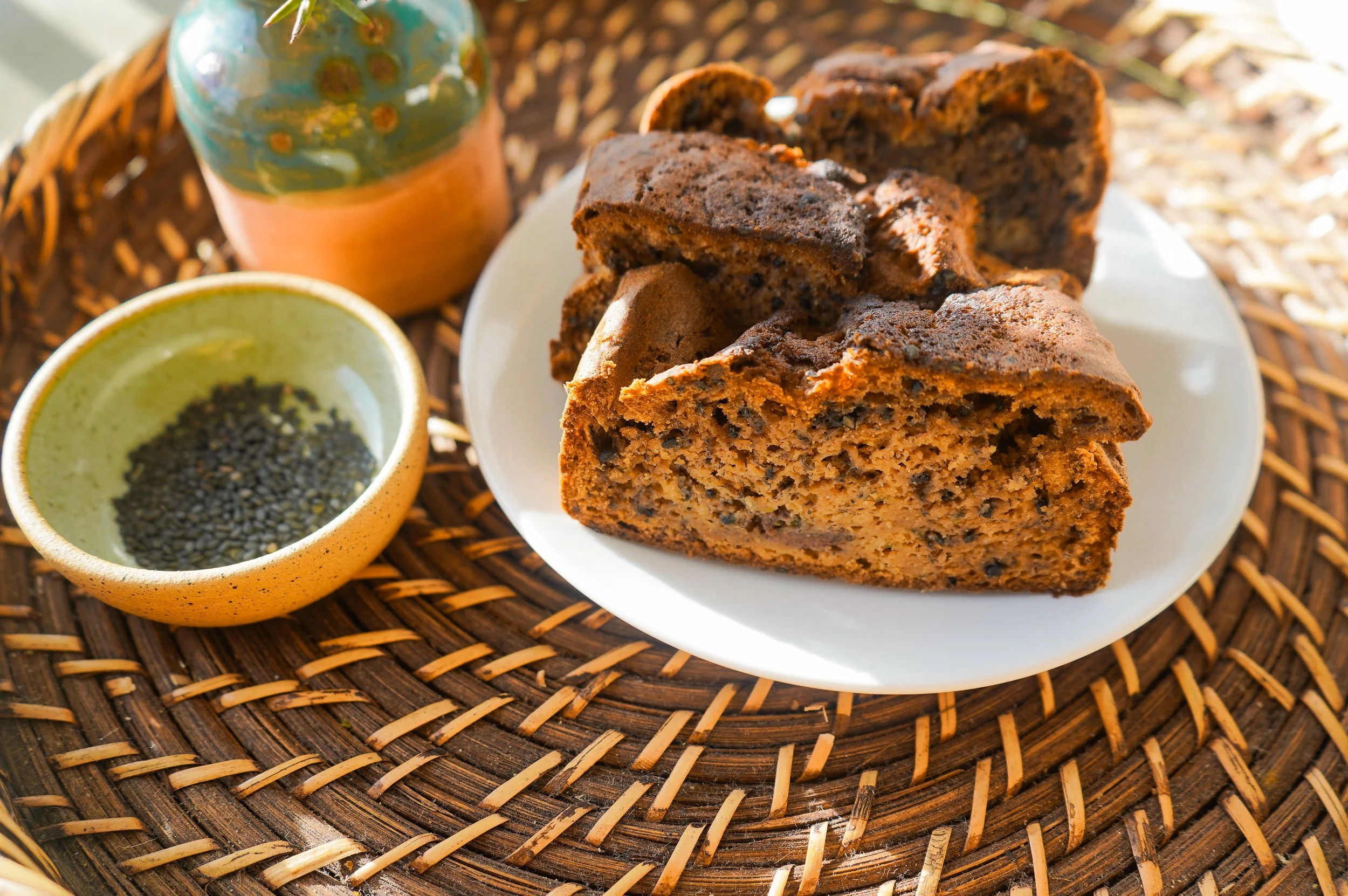 Slices of zucchini bread from The Delicate Squash on a white plate, a small bowl of black sesame seeds, and a rustic vase on a woven rattan placemat.