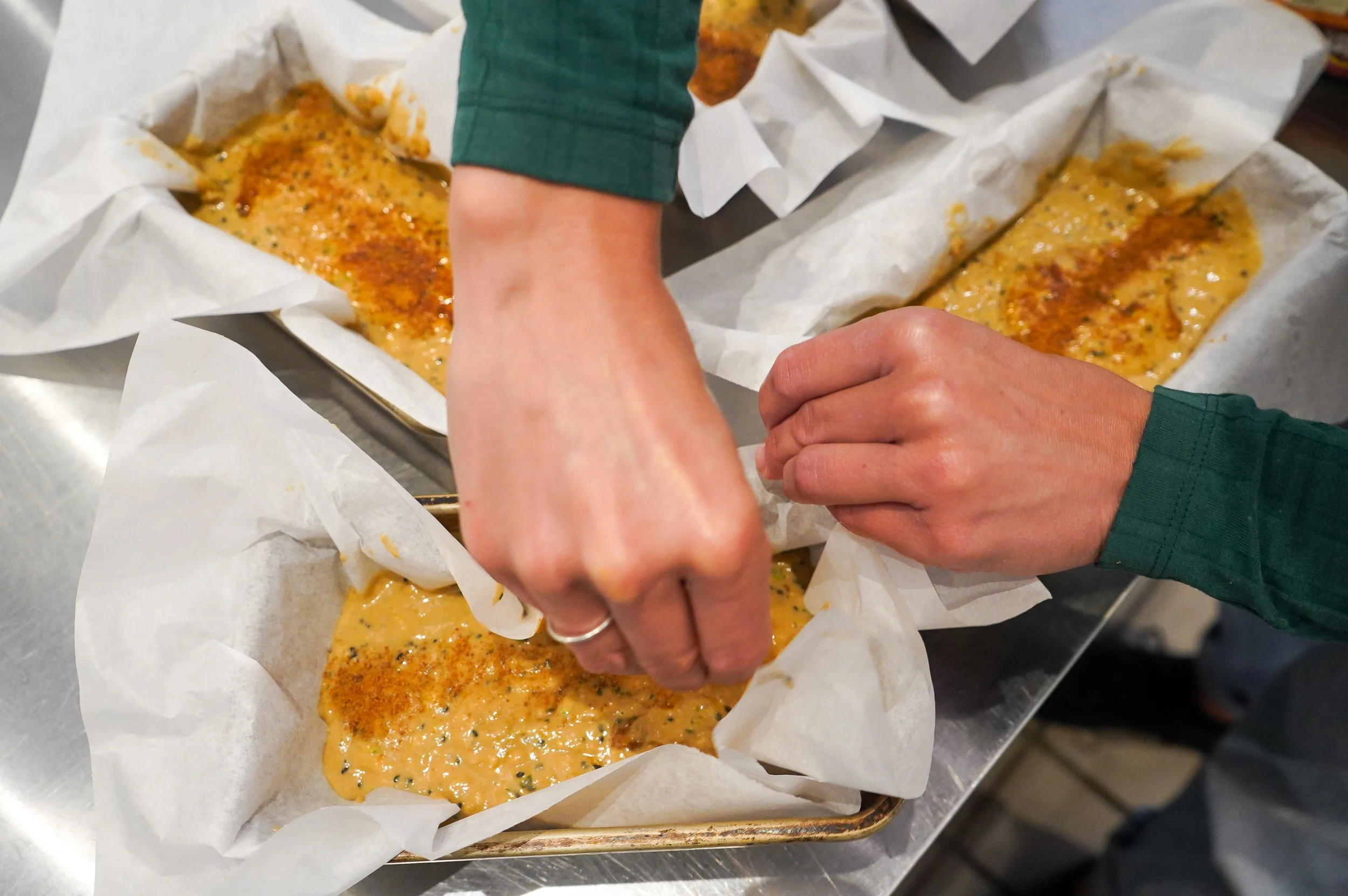 Baker from The Delicate Squash wearing a green long sleeve shirt preparing spiced banana bread. Batter with a line of sprinkled spices, is poured on parchment paper-lined trays for baking.