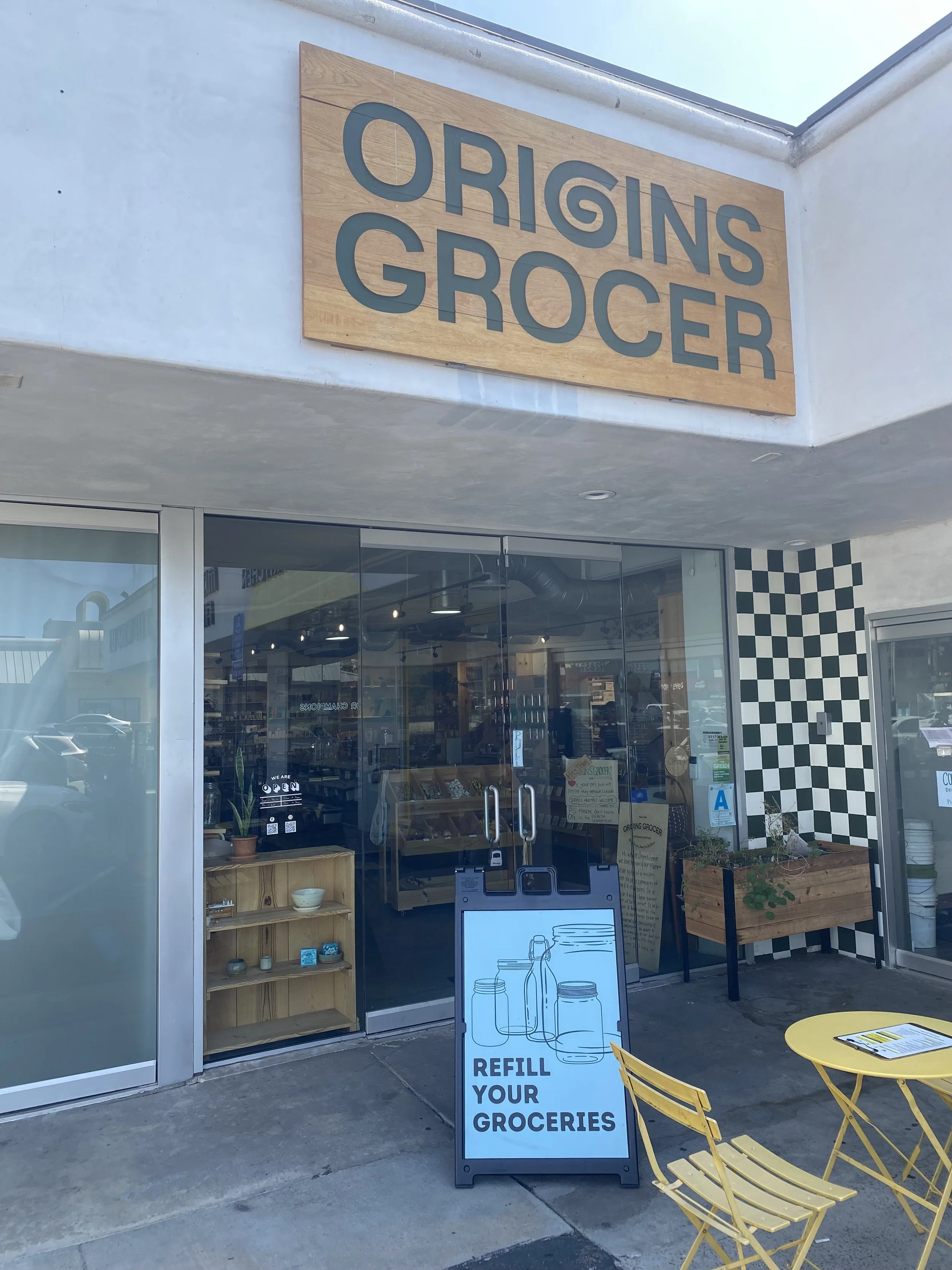 Facade of Origins Grocer with a wooden sign displaying the store's name. Glass doors lead inside where shelves and displays are visible. There is a white sandwich board sign that reads 'Refill Your Groceries' with illustrations of jars and bottles. Outside, yellow chairs and a small table are set up.