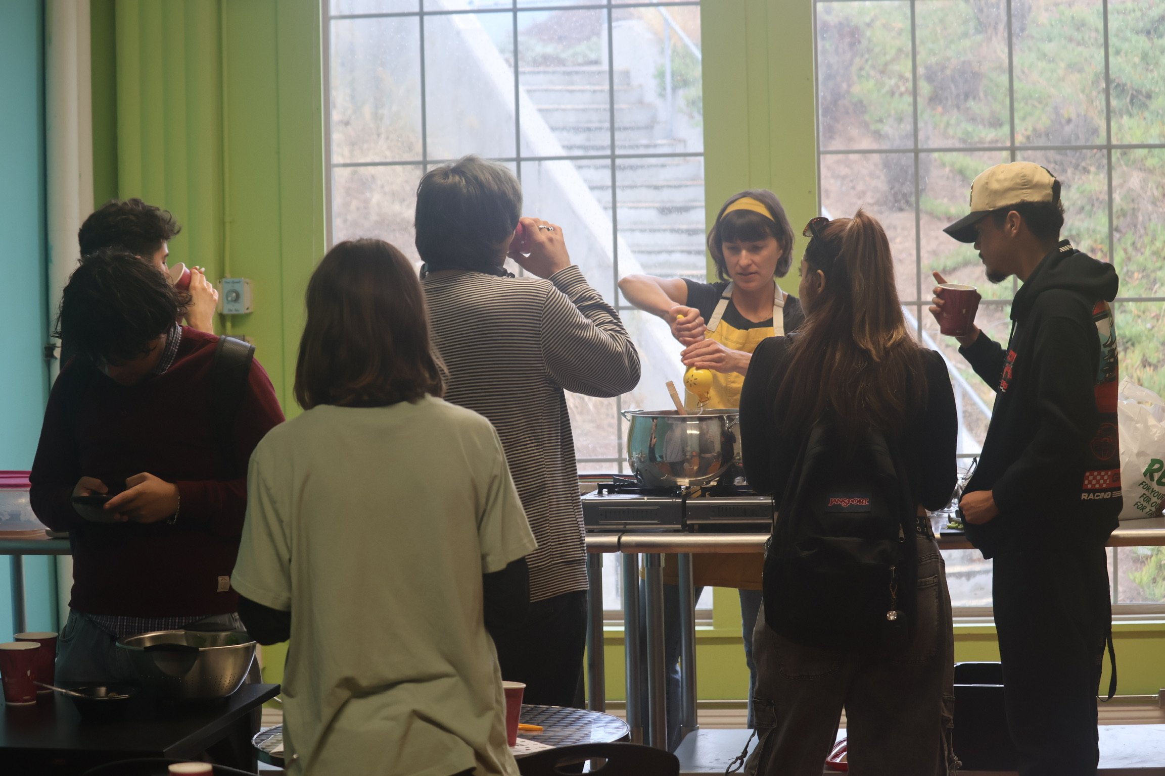 Group of people watching a woman cook in a kitchen with large windows.