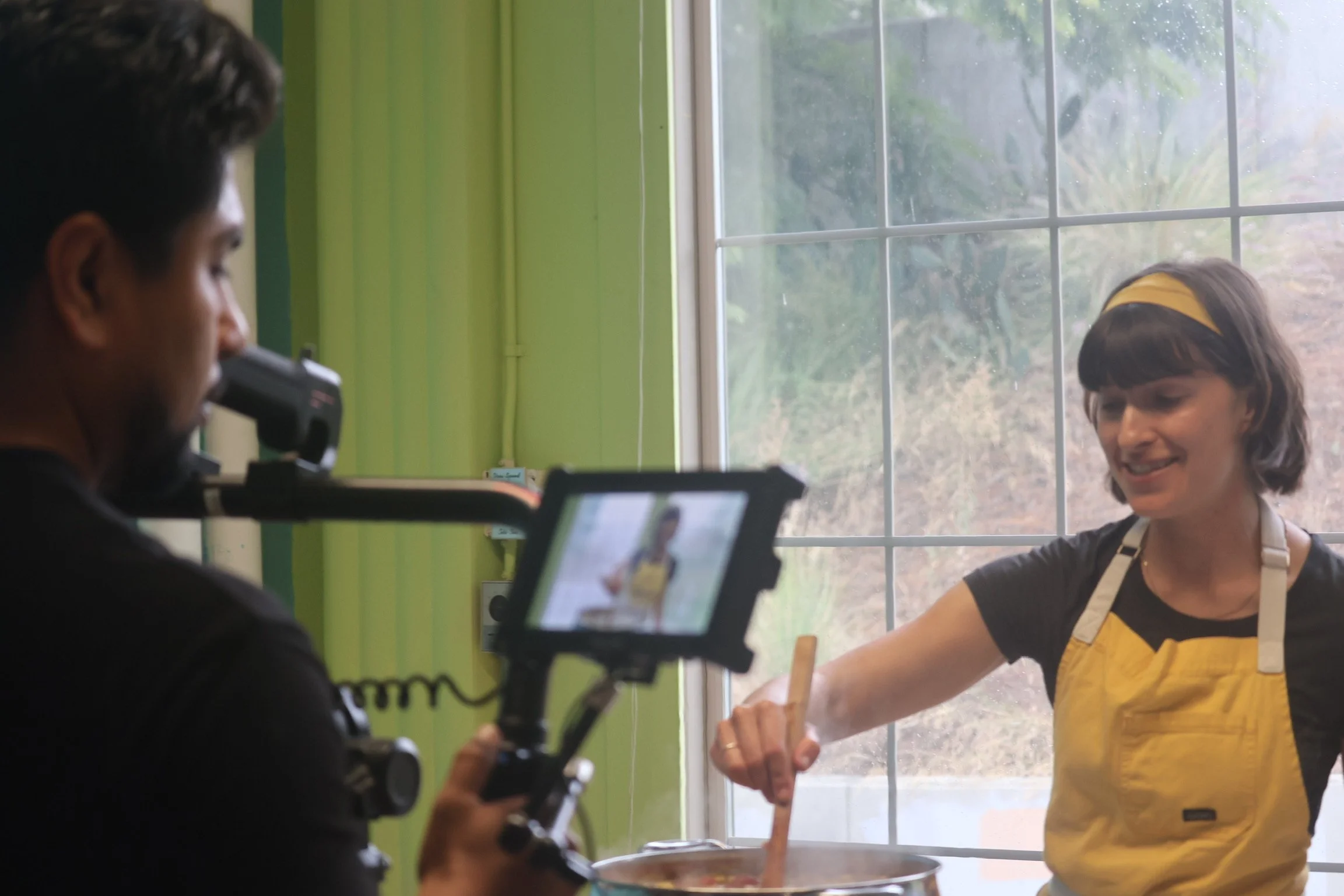A woman in a yellow apron and headband stirring a pot while being filmed by a person holding a camera mounted on a stabilizer inside a green-walled kitchen with a large window.
