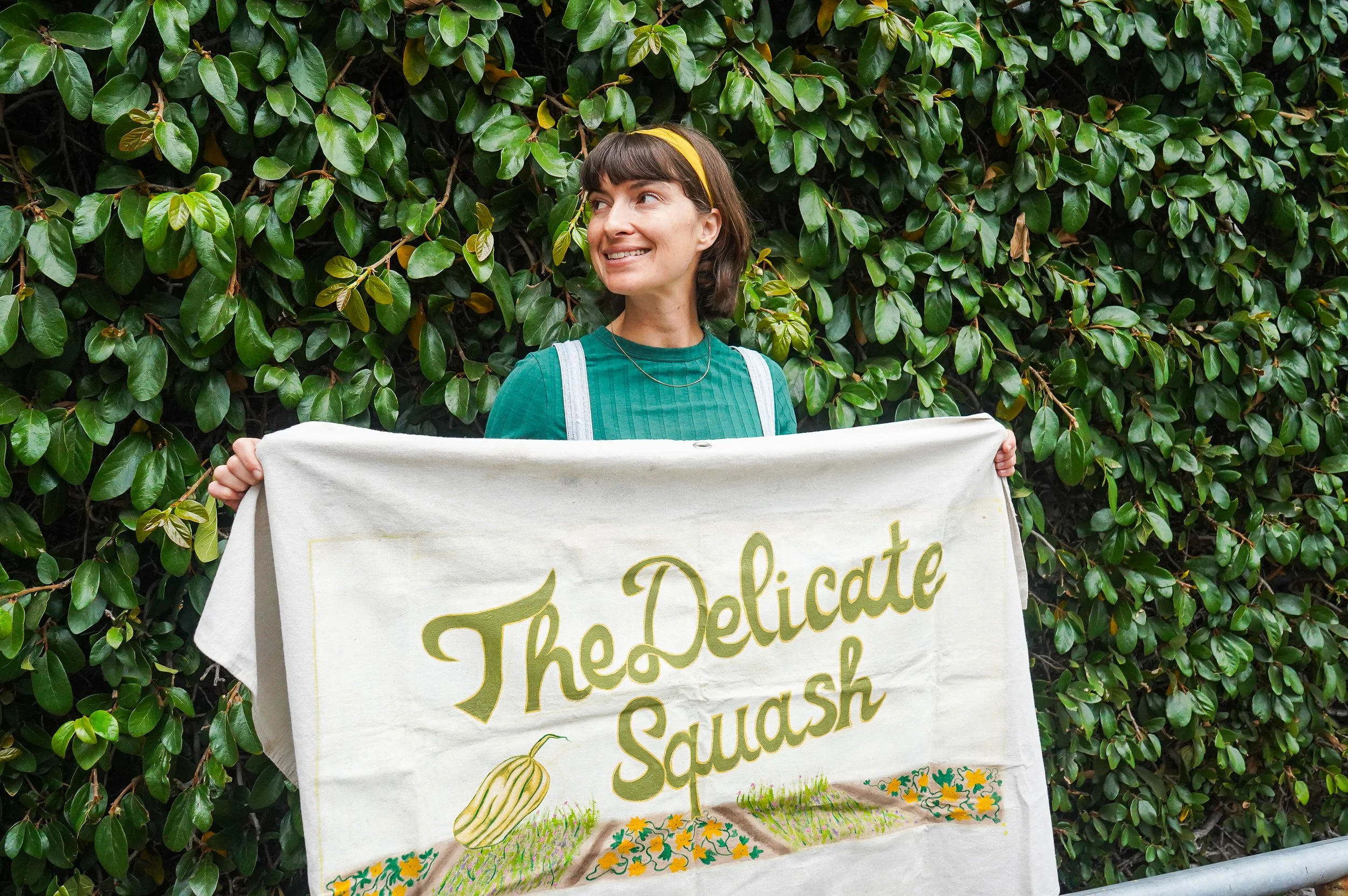 A woman holding a banner that reads 'The Delicate Squash' in front of a dense green leafy hedge.