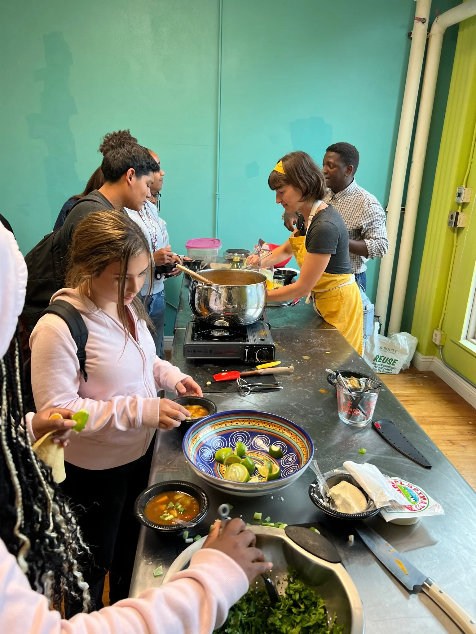 Group of people cooking and serving food in a kitchen or cooking class setting with various ingredients and utensils on the counter.