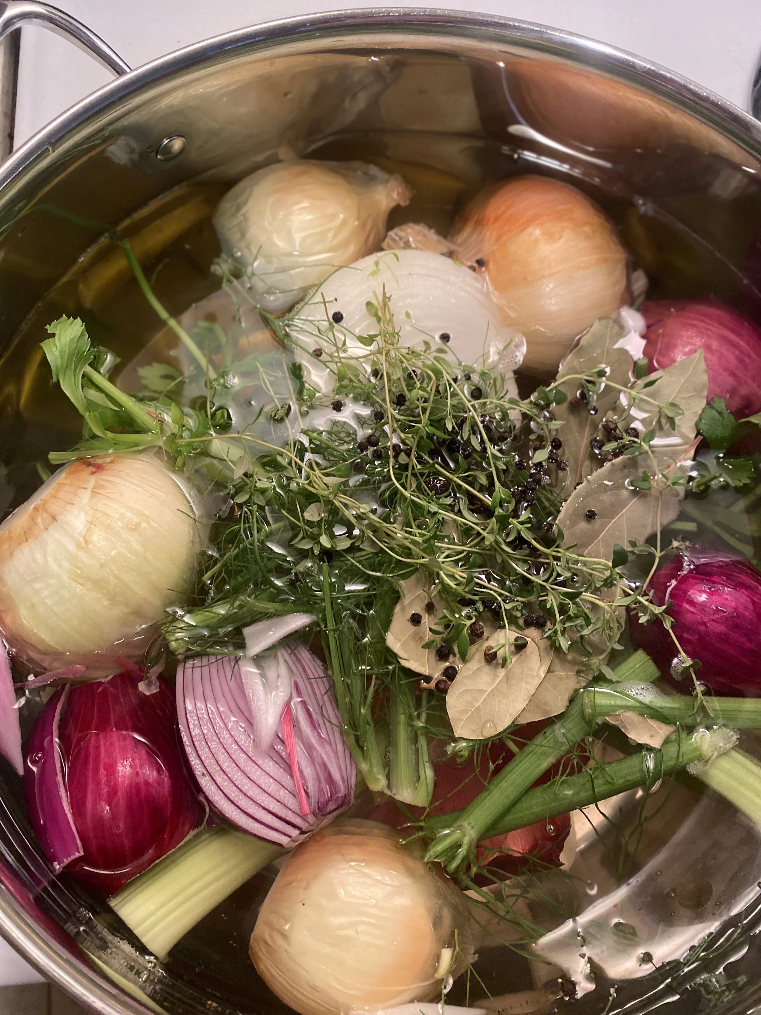A pot filled with water containing onions, garlic, shallots, bay leaves, black peppercorns, celery stalks, and sprigs of fresh thyme and other herbs for making broth or stock.
