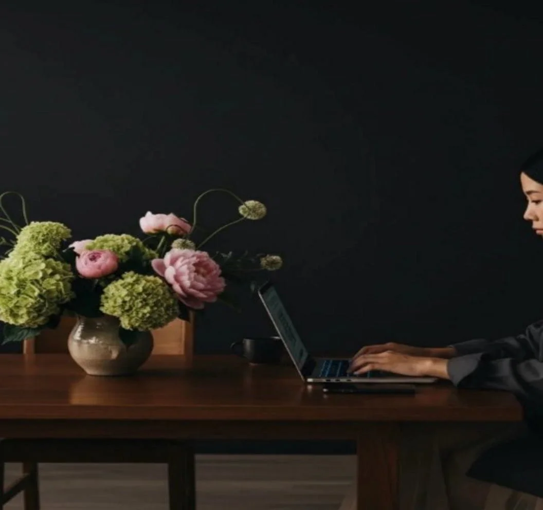 A woman working on a laptop at a wooden table with a vase of pink and green flowers against a dark wall.