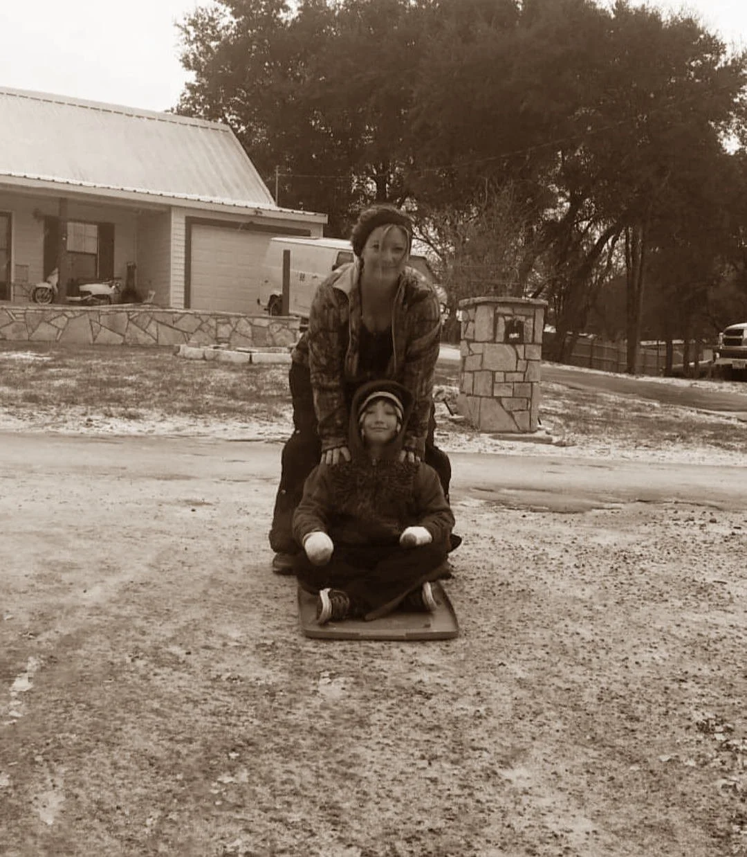 A woman and a young boy are playing outside on a sled. The woman is standing behind the boy, smiling, while the boy is sitting on the sled with a relaxed expression. The scene appears wintry, with some snow on the ground and trees in the background.