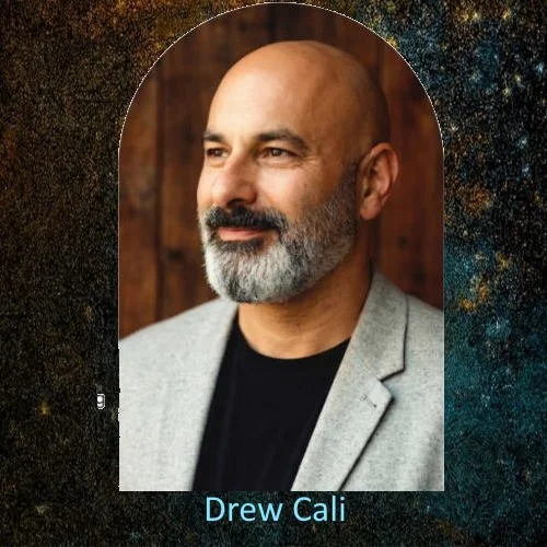 Photograph of Drew Cali with a bald head and gray beard, wearing a light gray blazer and black shirt, standing against a wood-paneled background.