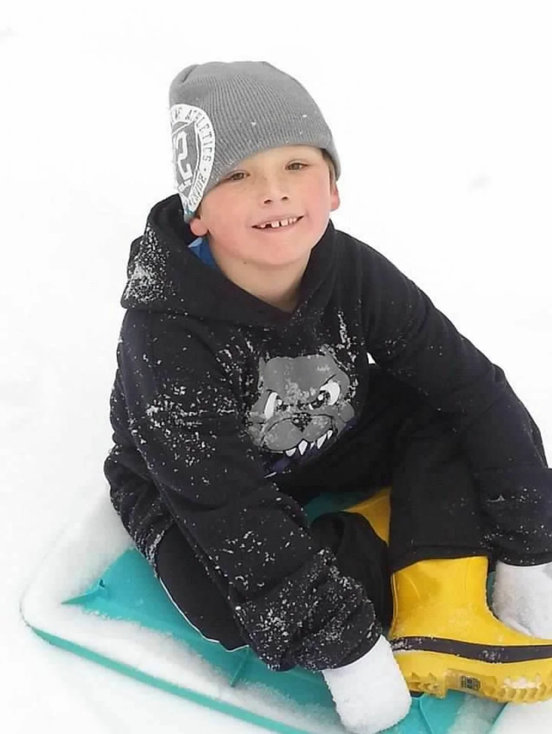 A young boy with a gray beanie smiling while sitting on a yellow sled in the snow.