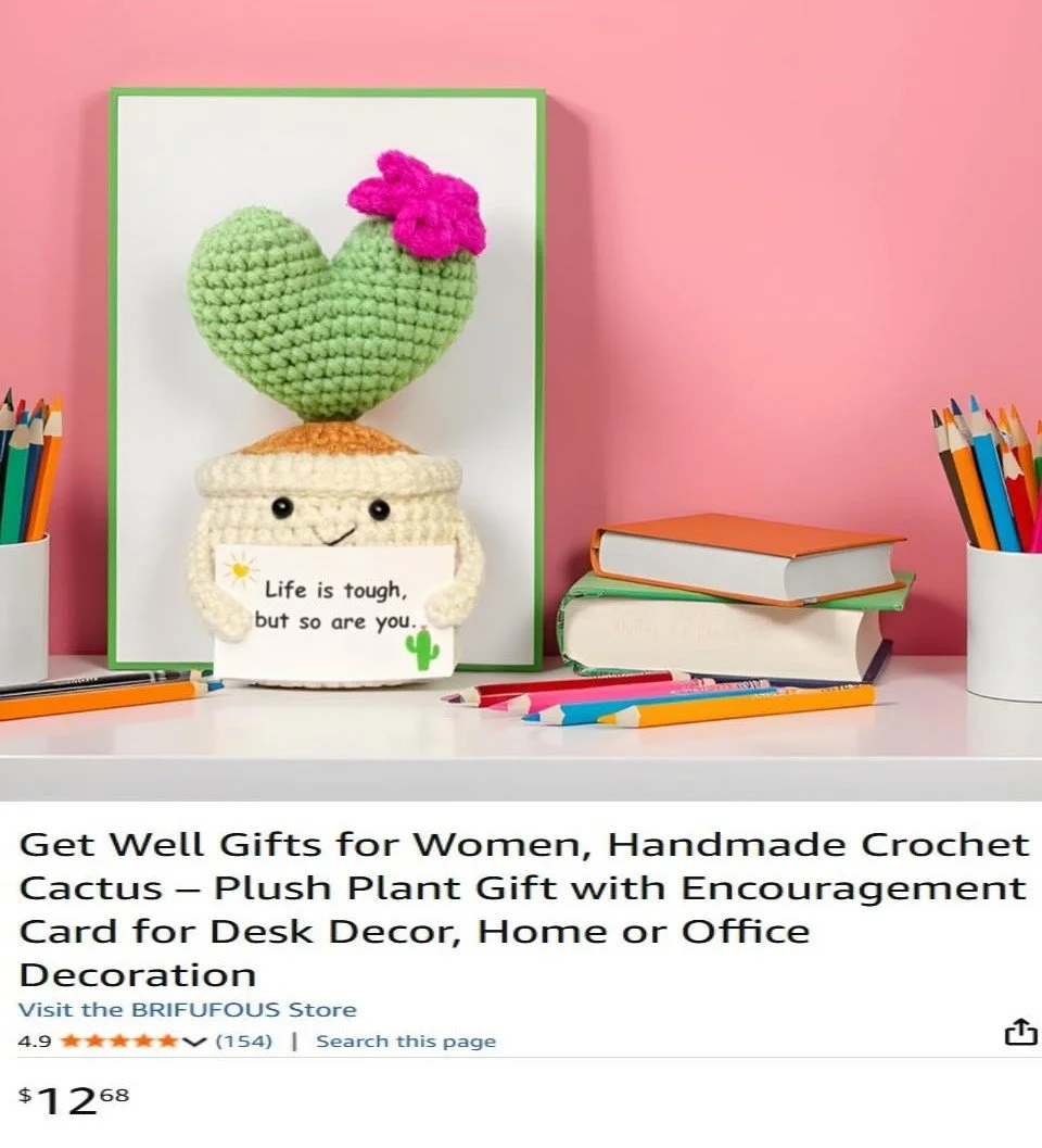 A crochet plush cactus with a pink flower on top, holding a small sign that reads "Life is tough, but so are you". The cactus is in a crochet pot with a smiley face, placed on a white desk with colorful pencils and notebooks.