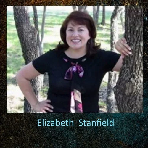 A woman with shoulder-length dark hair, smiling, wearing a black shirt with a purple bow, standing outdoors among trees.