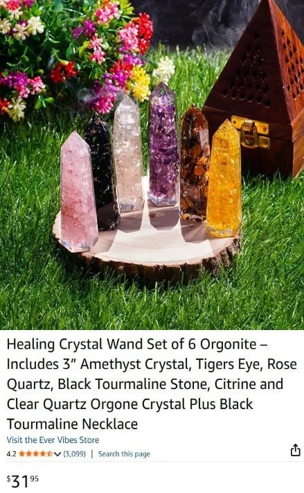 Set of six healing crystal wands with various colors and types, displayed on a wooden disk outdoors with green grass, flowers, and a wooden pyramid-shaped box in the background.