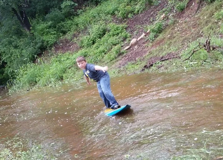 A boy is riding a skateboard on a shallow stream in a wooded area.