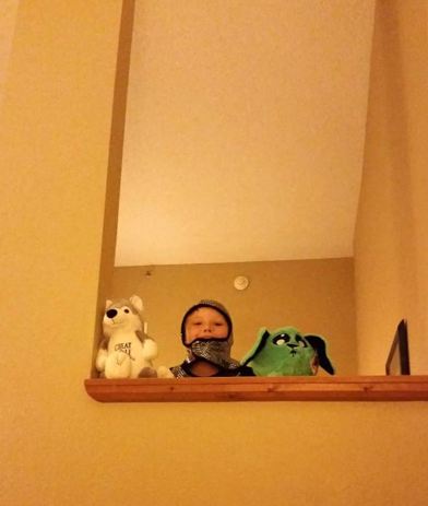 A young boy standing at a kitchen or balcony bar, looking over the edge, with a stuffed white dog and a green plush toy on the counter behind him.