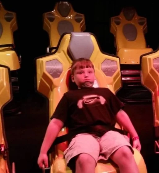 A young boy sitting in a yellow simulated ride or chair with other similar chairs in the background.