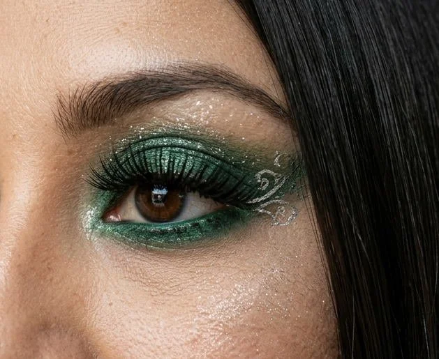 Close-up of a woman's eye with green and silver glitter eyeshadow, black eyeliner, voluminous eyelashes, and decorative silver swirl details near the outer corner.