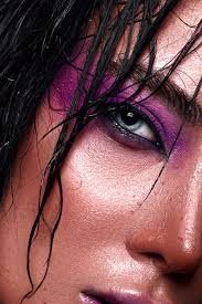 A close-up of a woman's face, showing her eye with purple eyeshadow, wet black hair, and shiny skin.