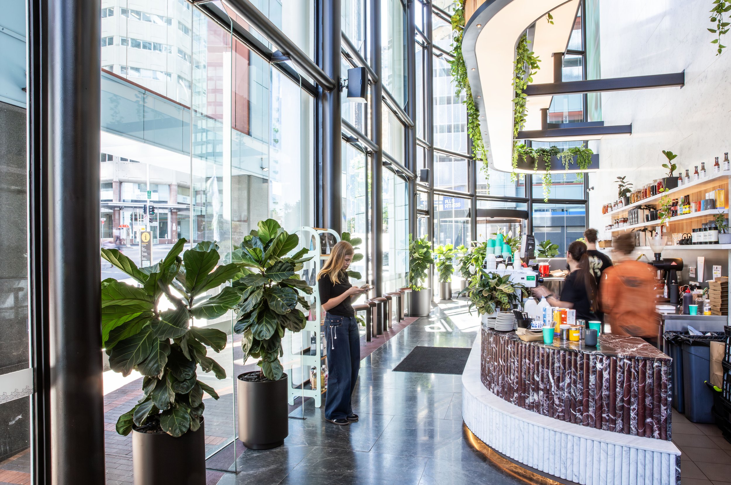 Inside a modern, sunny coffee shop with large glass windows, green plants, and a marble counter. Customers and staff are present, with one woman standing near the entrance looking at her phone.