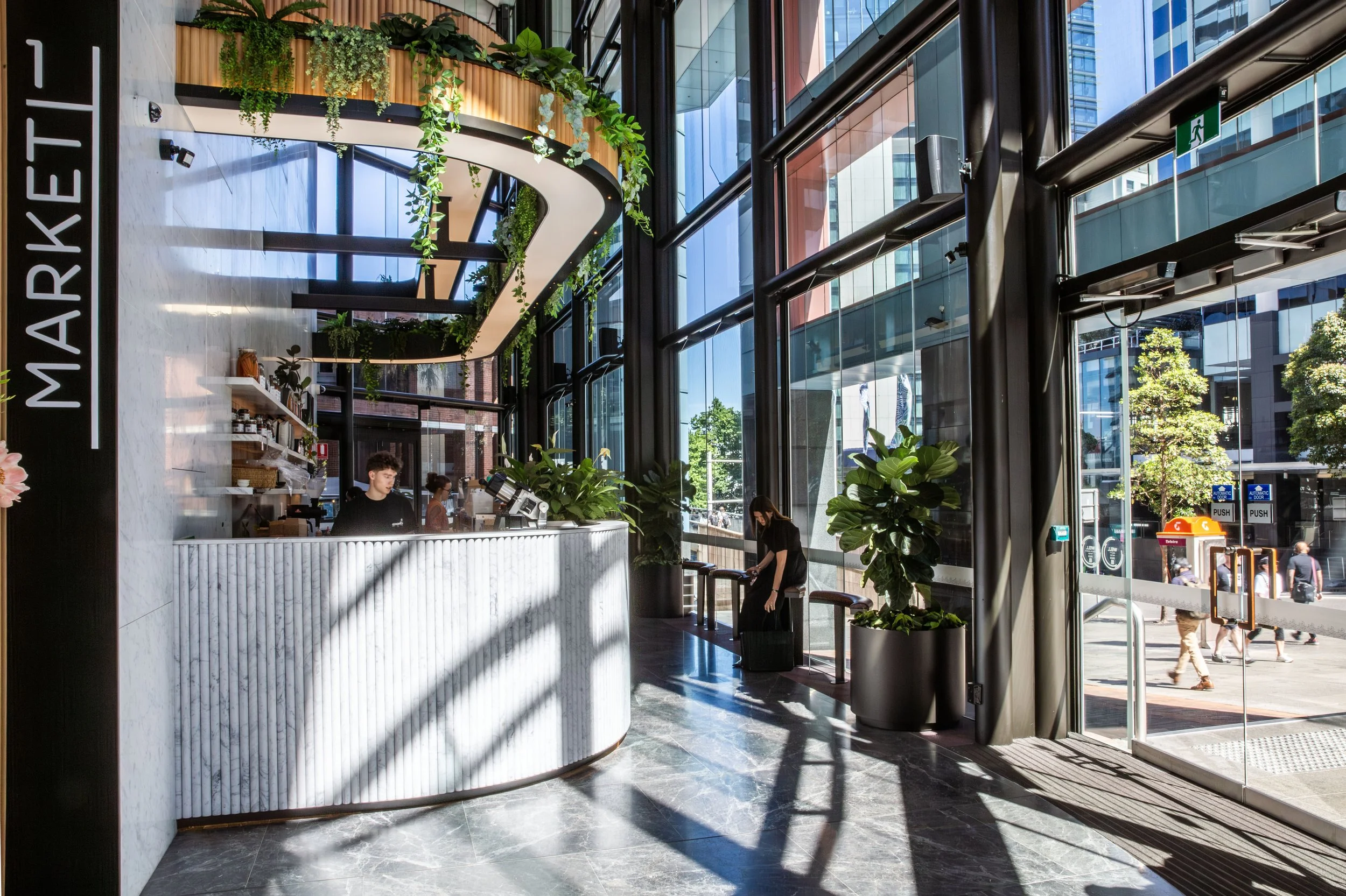 Inside a modern, well-lit marketplace with large glass windows, a curved white marble counter, and green plants. Two people are near the counter, one working and the other tending to something. Outdoors, people are walking on the sidewalk, with trees and buildings visible through the windows.