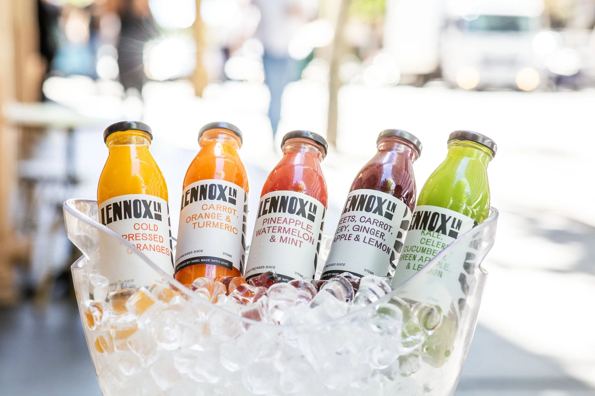 Five bottles of fresh fruit and vegetable juices in a metal container filled with ice, with a blurred outdoor background.