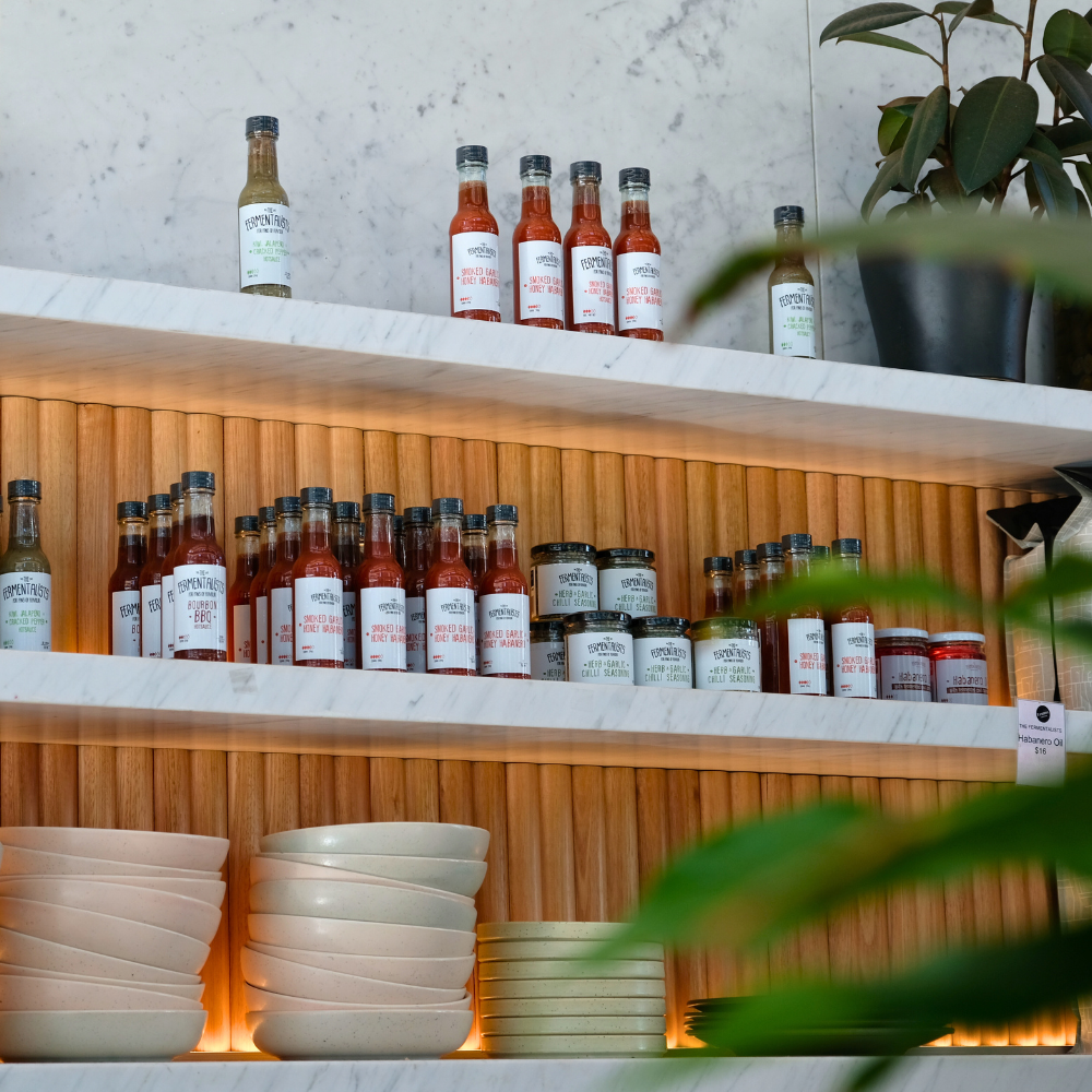 Shelves with bottles of hot sauces and jars, and stacked bowls and plates, with a potted plant on the side.
