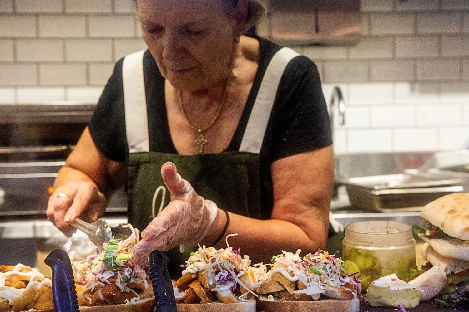 An elderly woman with purple hair, wearing a black shirt and a green apron, is preparing food in a kitchen. She is using tongs to add toppings to a dish that appears to be tacos, which are topped with shredded vegetables and sauces. There are other sandwiches and ingredients on the counter, and a jar of pickles or relish is visible.