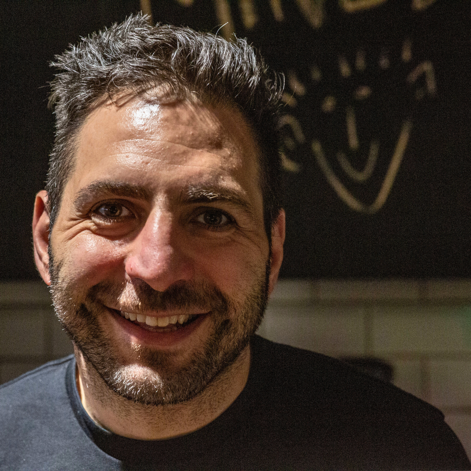 A man smiling with a dark background and some indistinct writing or graffiti behind him.