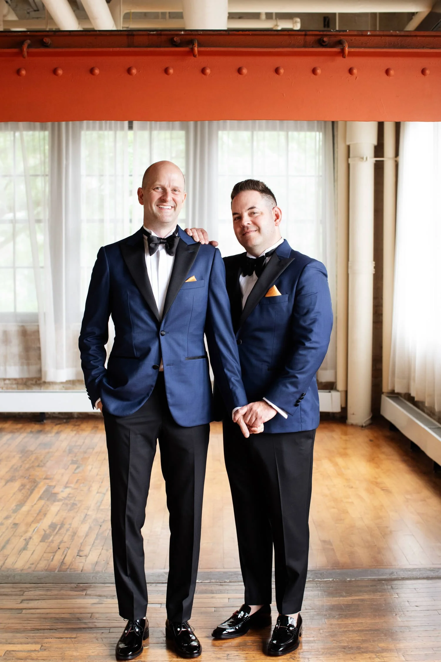 The two grooms in classic black and navy blue suits and black bow ties and details at their Minneapolis wedding at Machine Shop. Planned by Rocket Science Events