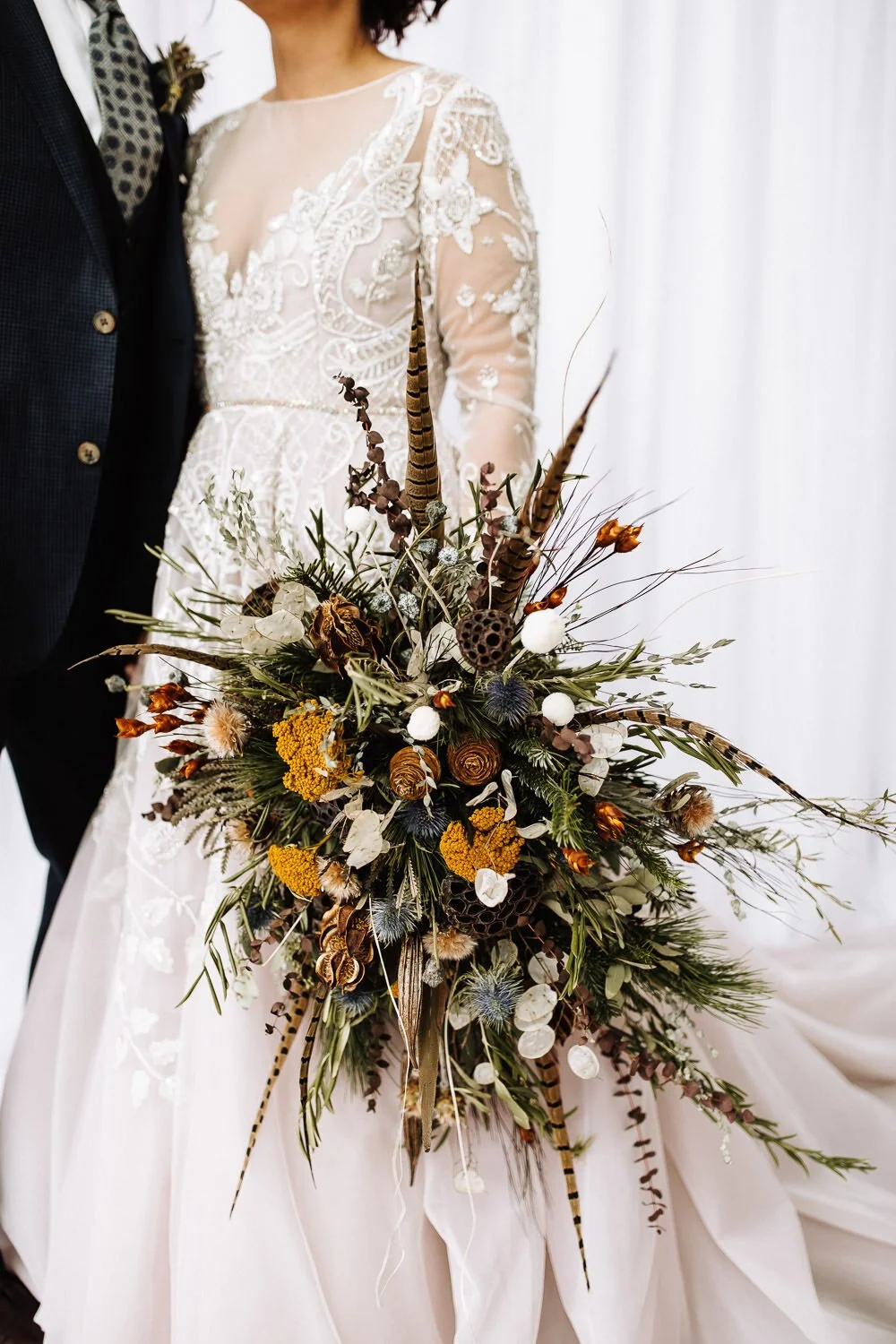 bride in long sleeved lace gown holding modern large bouquet of dried flowers and feathers
