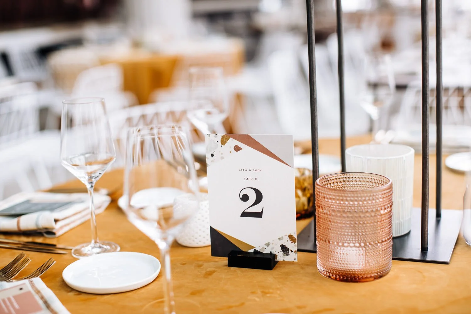 table number, geometric design with mustard yellow table cloth, dusty pink glass votive candle holder, and long-stemmed wine glasses.
