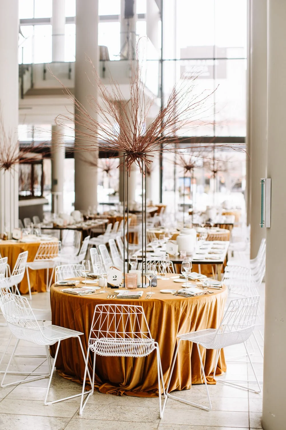 wedding tablescape of a round table with mustard yellow velvet table cloth, the tall centerpiece of burnt orange twigs, and modern white wire eames chairs.