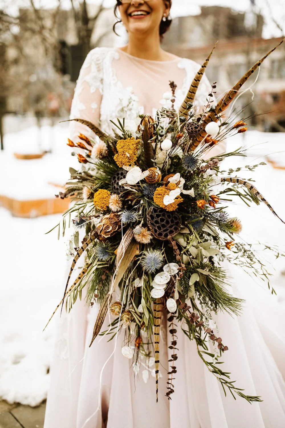 bride in long-sleeved lace gown holding a modern large bouquet of dried flowers and feathers, smiling outside in the snow