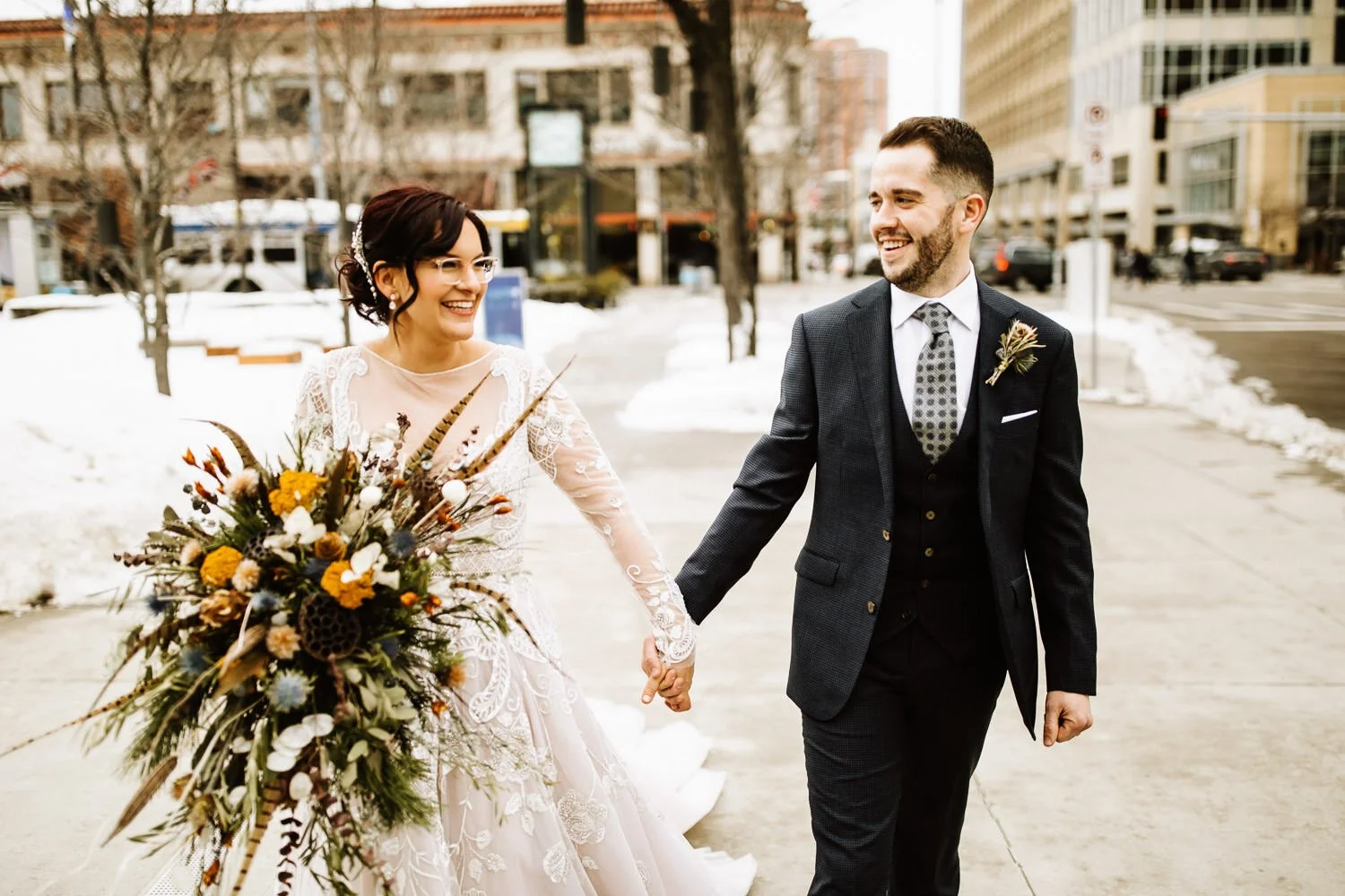bride wearing long-sleeved lace gown holding hands and smiling with the groom wearing a dark charcoal grey suit. bouquet of dried flowers and feathers