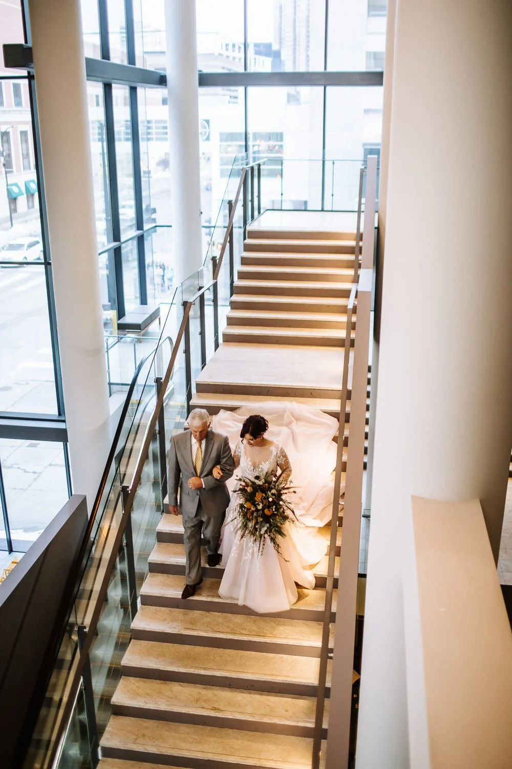 father walking bride downstairs in modern glass museum setting with long veil trailing behind