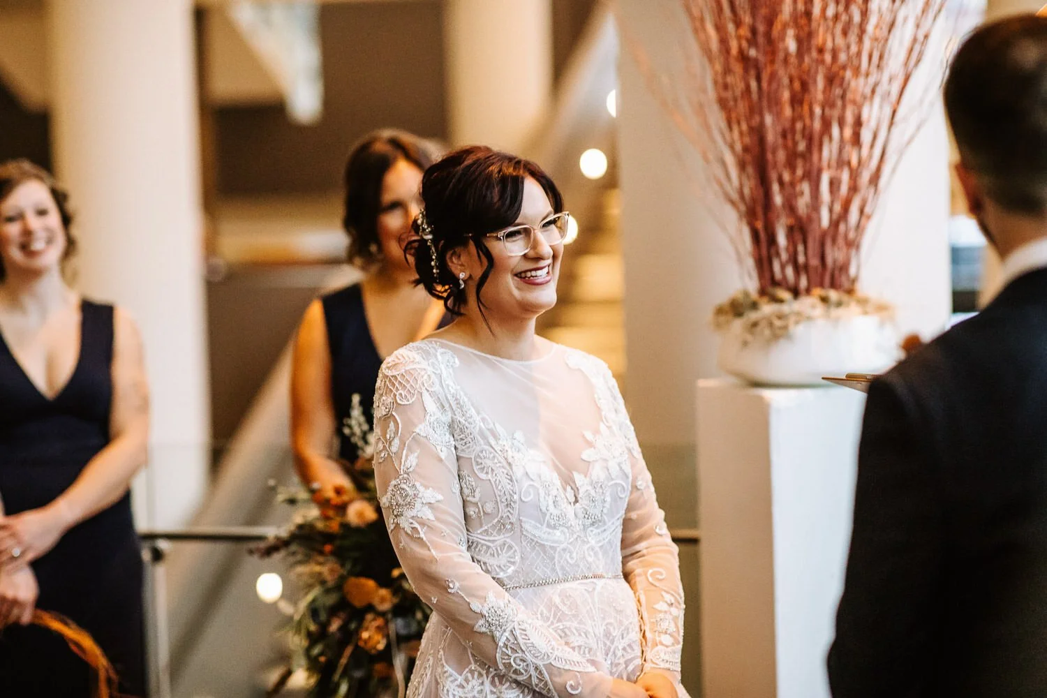 bride in long-sleeved lace gown and glasses smiling during ceremony