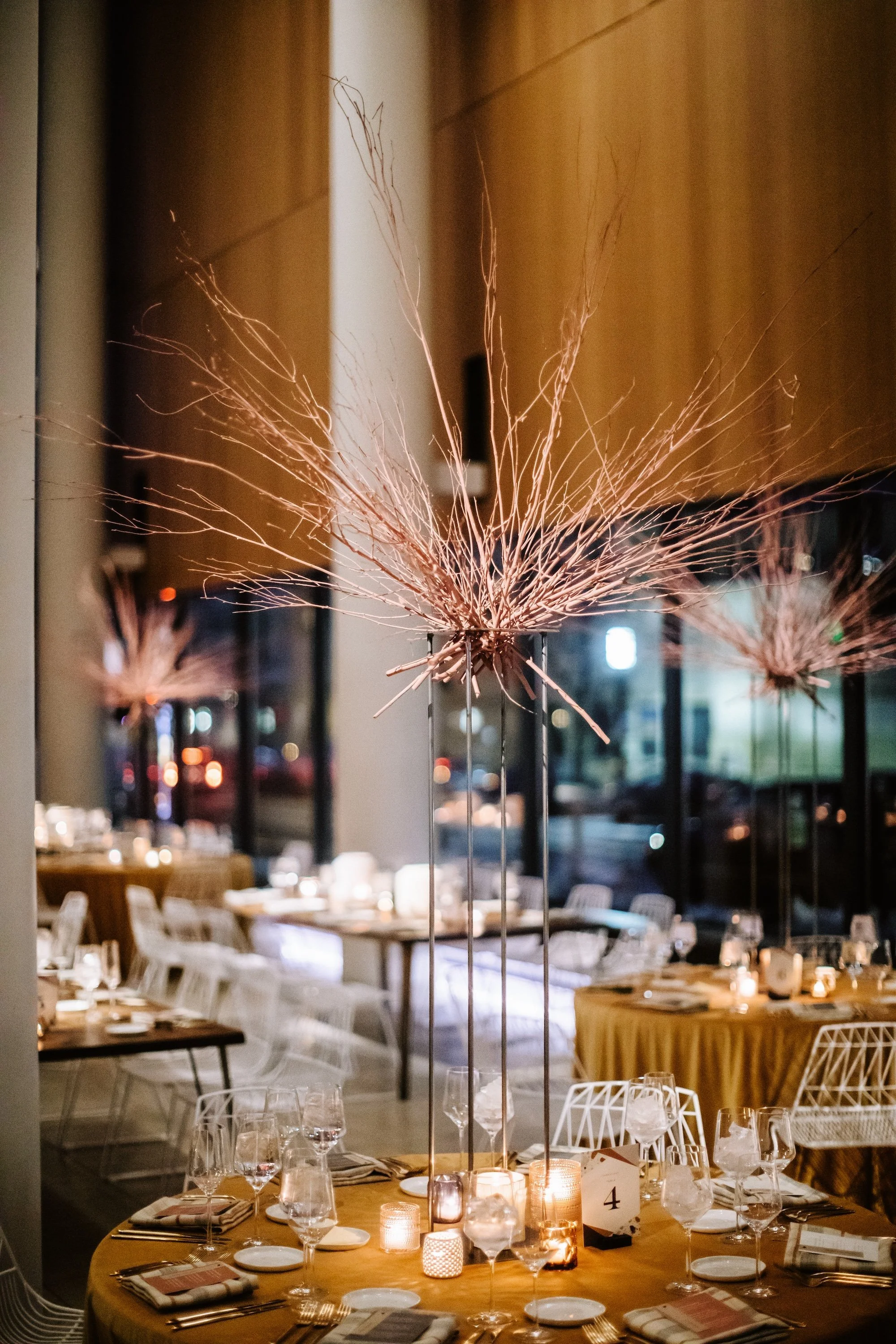 wedding tablescape of a round table with mustard yellow velvet table cloth, the tall centerpiece of burnt orange twigs, and modern white wire eames chairs.