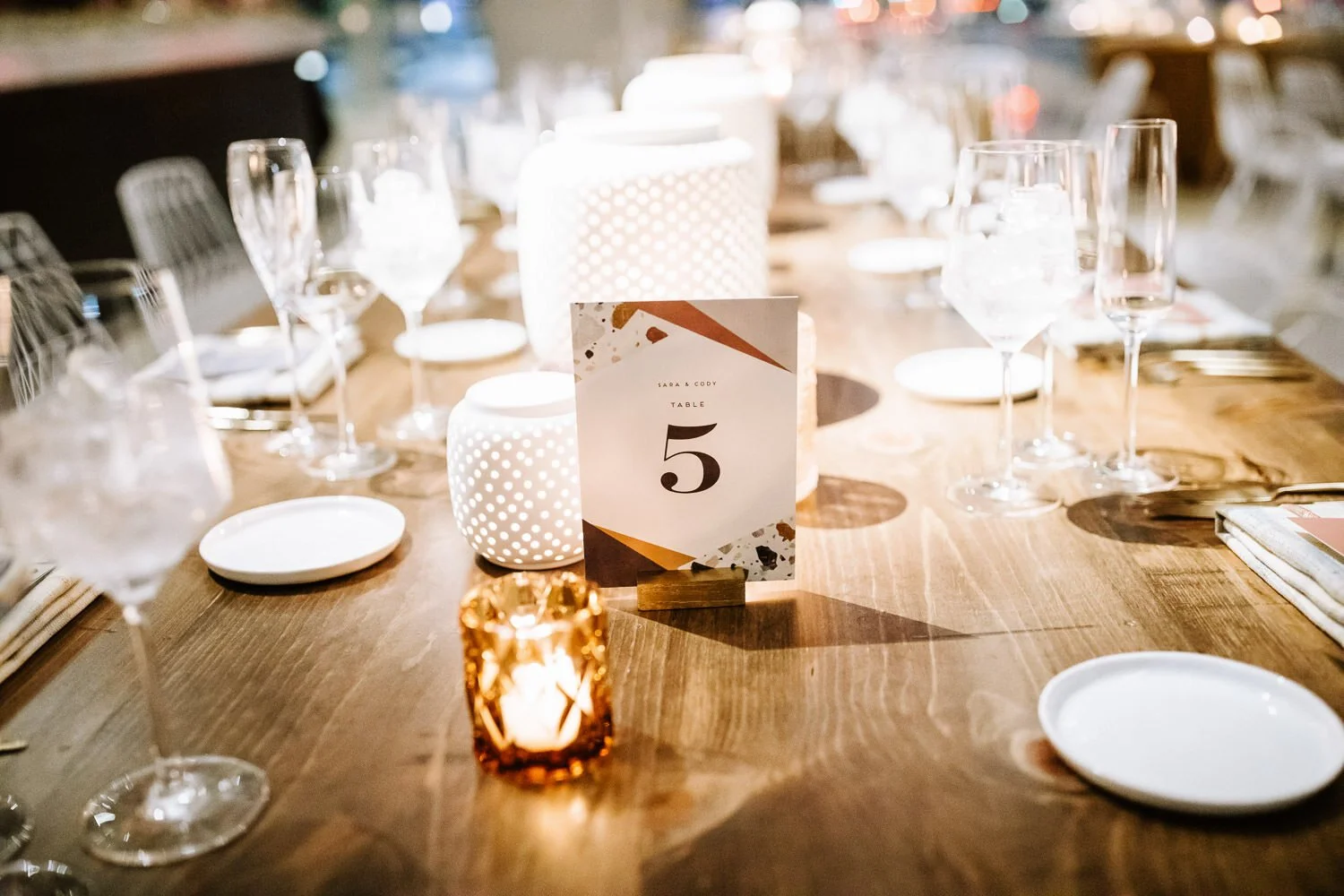table number, geometric design on wooden farm table, dusty pink glass votive candle holder, and long-stemmed wine glasses.