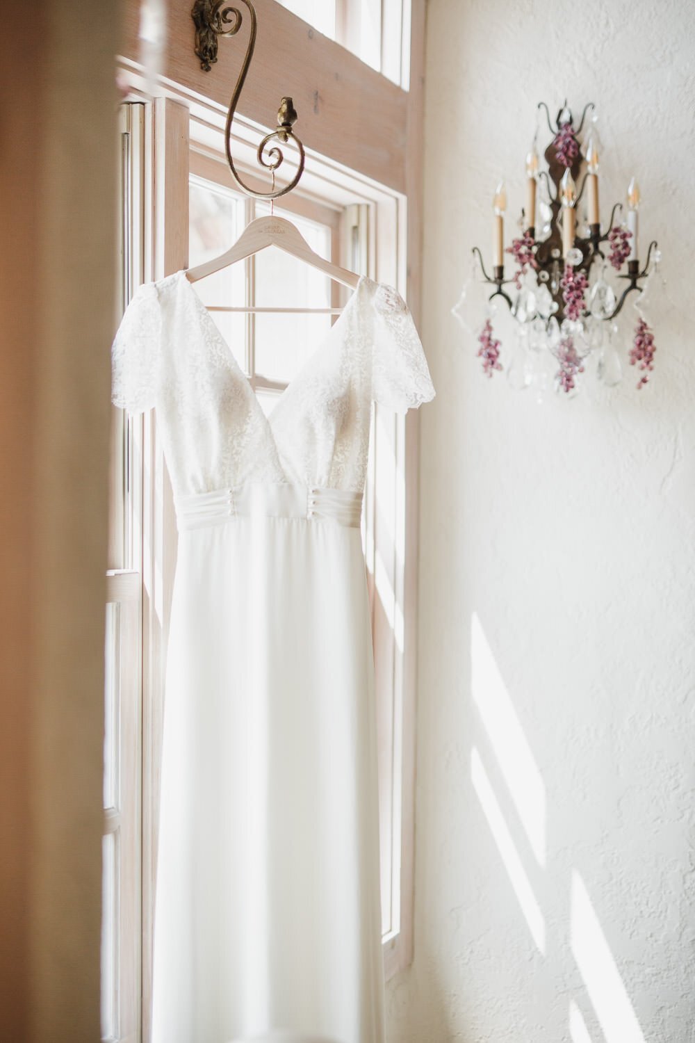 wedding dress with lace cap sleeves, v neck and silk bodice hanging in the sunlit window with wall sconce - Rocket Science Events